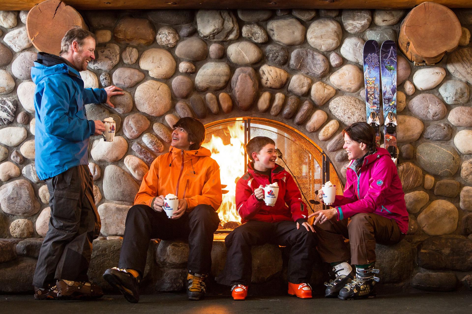 Parents and children enjoy warm drinks by a fireplace after skiing