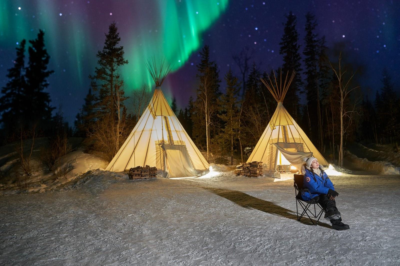 Yuna Kim sits outside two teepees at night, surveying the Northern Lights