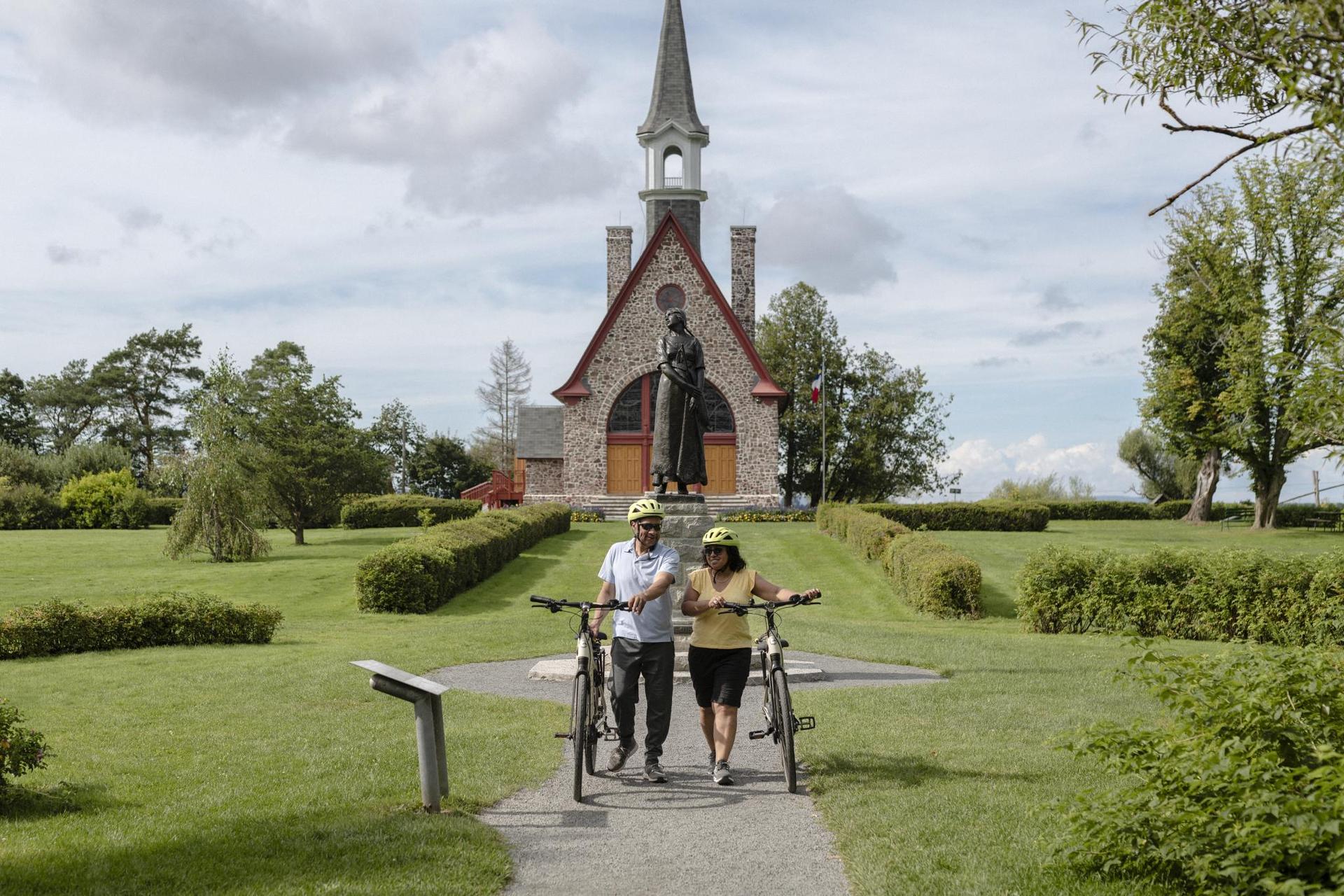 Two people walk away with bikes from a historic site - a green grassy area with a church. 