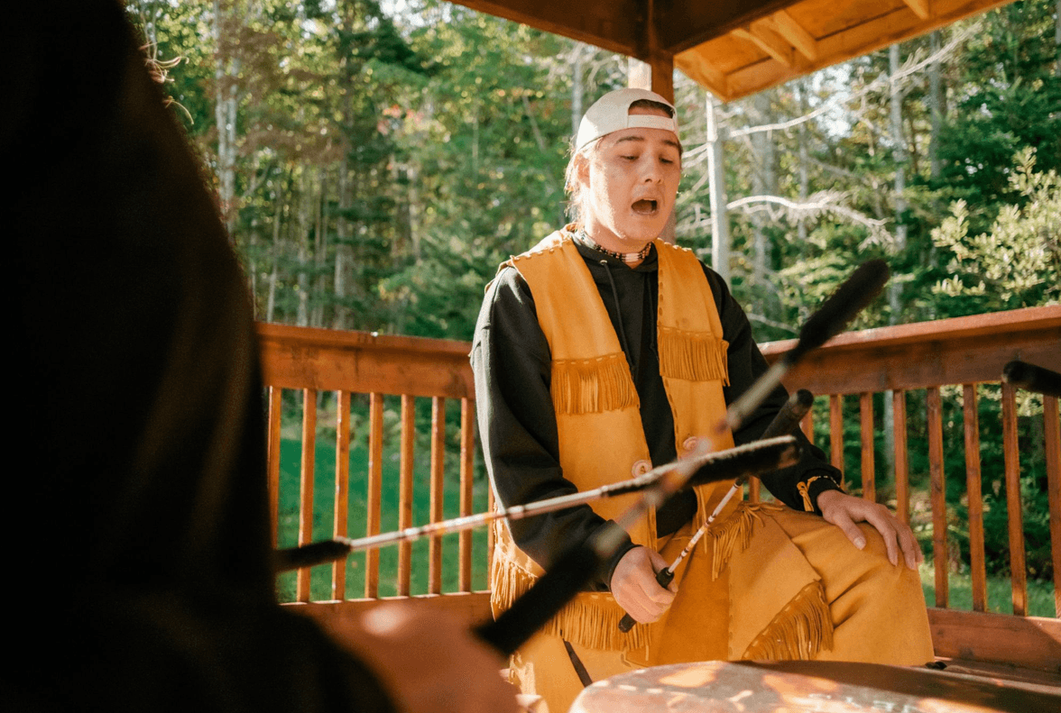 Indigenous person sings on porch with forest in background in a drum circle