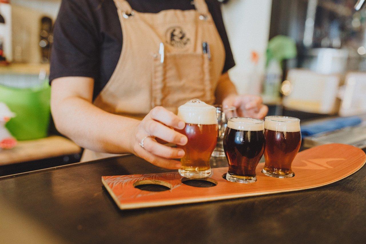 Person in apron with a flight of different coloured beers on a wood holder handing one towards camera.