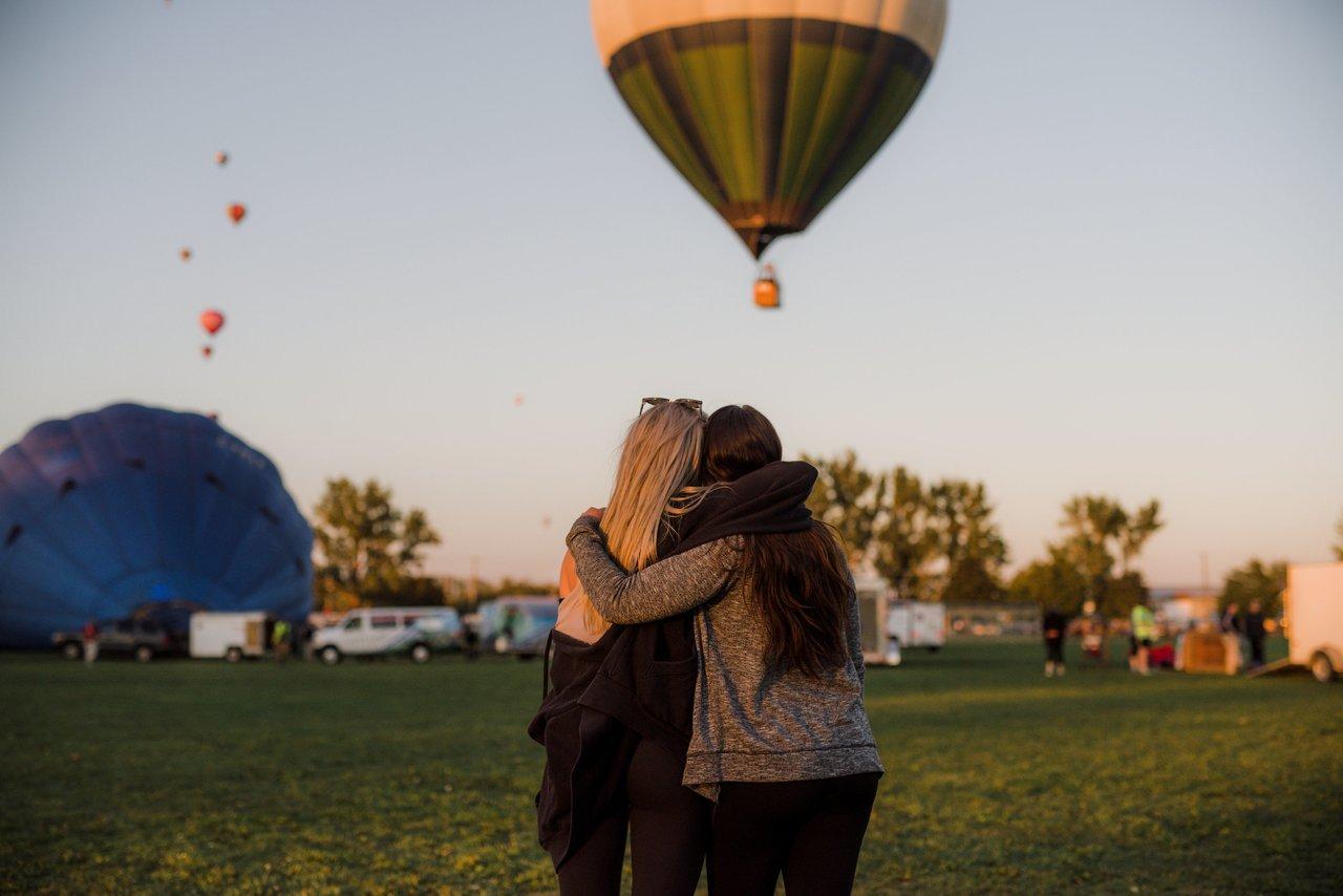 Two people hold each other on a grassy area watching hot air balloons float to the sky