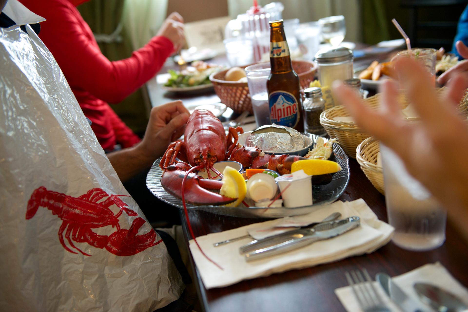 Lobster appears on a table surrounded by people ready to enjoy with bibs and bread baskets. 