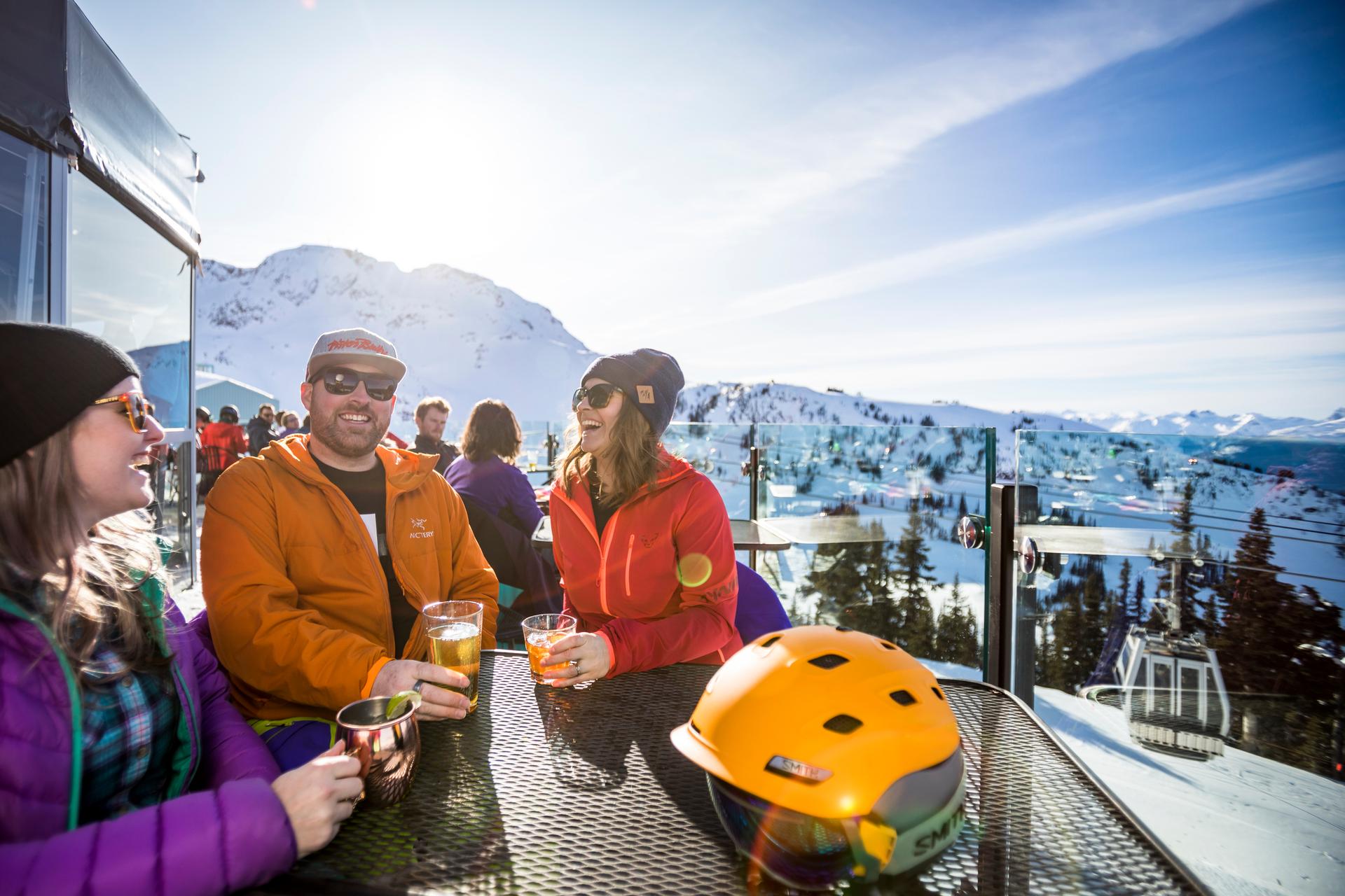 A group of friends enjoys drinks at a sunny mountain terrace with snow-covered slopes and ski lifts in the background.