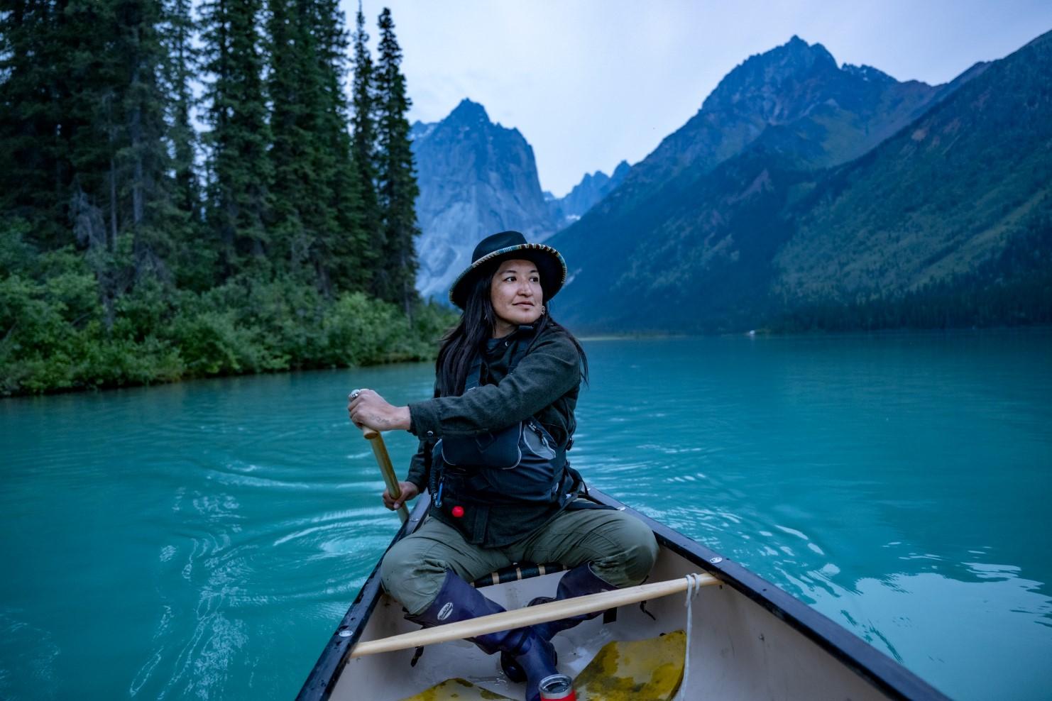 Person paddling a canoe on a calm turquoise lake, surrounded by mountains.