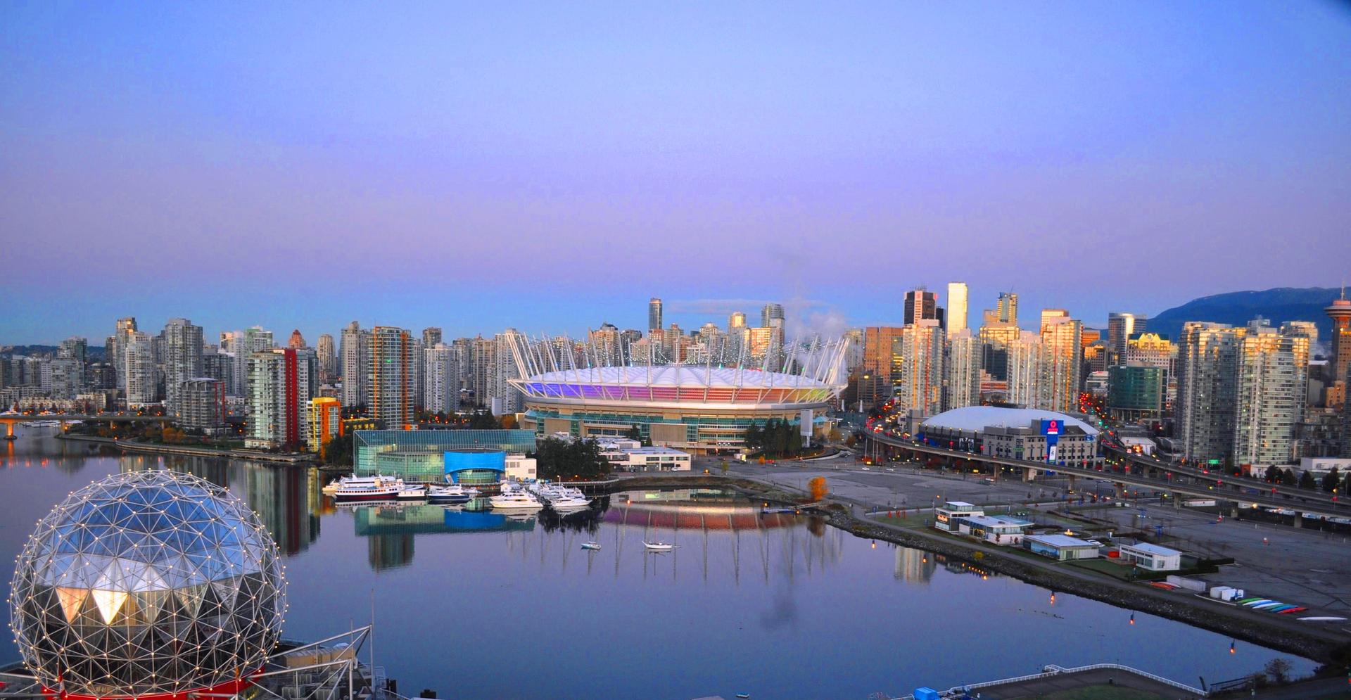 Vancouver skyline at sunset featuring BC Place, and a waterfront, reflecting in calm water.