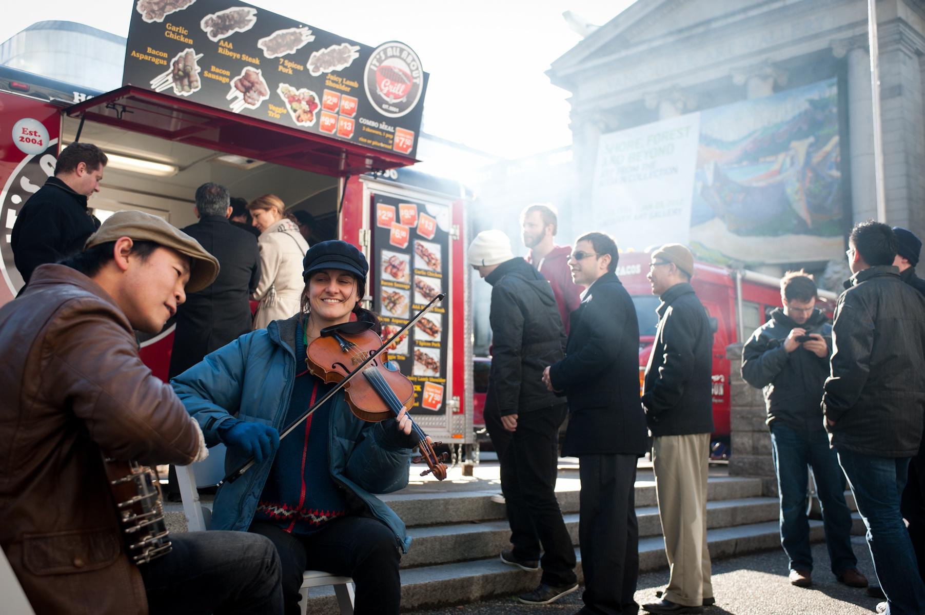 People stand in line at a food truck while musicians play instruments nearby, creating a lively street scene.