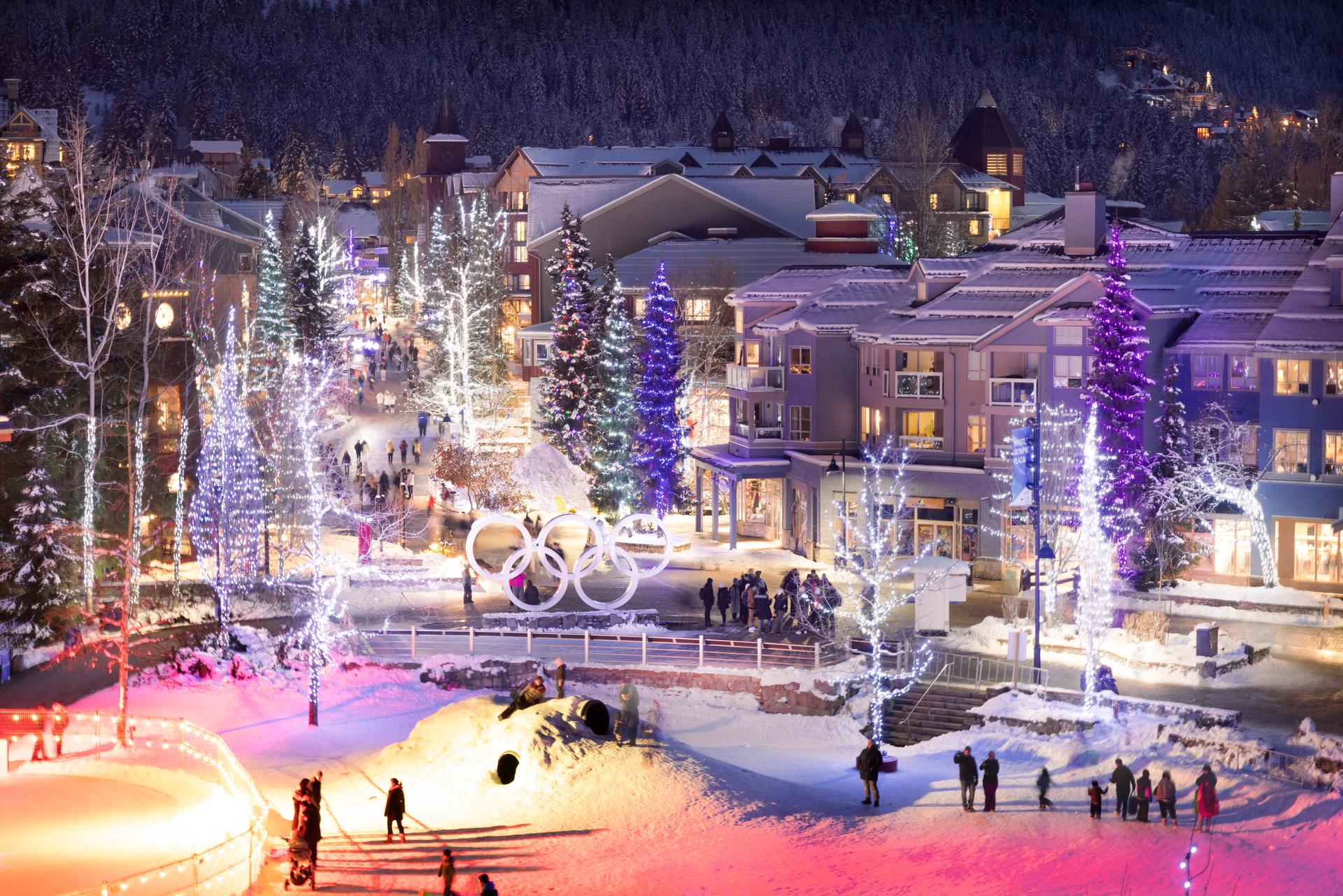 A lively Whistler village at night, decorated with colorful lights and lit-up Olympic rings.