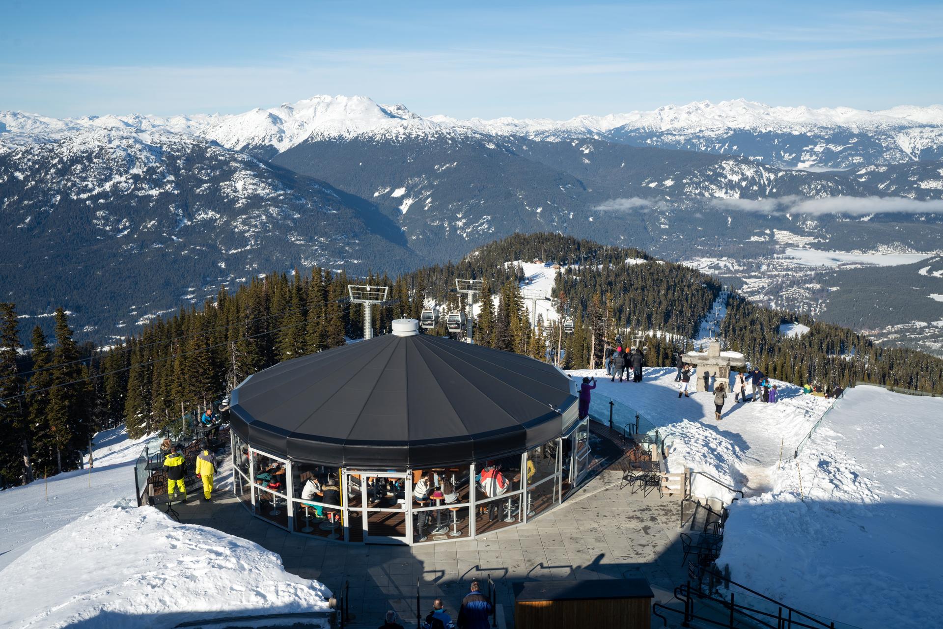 A circular mountaintop lodge sits surrounded by snow, with people enjoying the view of distant snowy mountains and forested slopes.