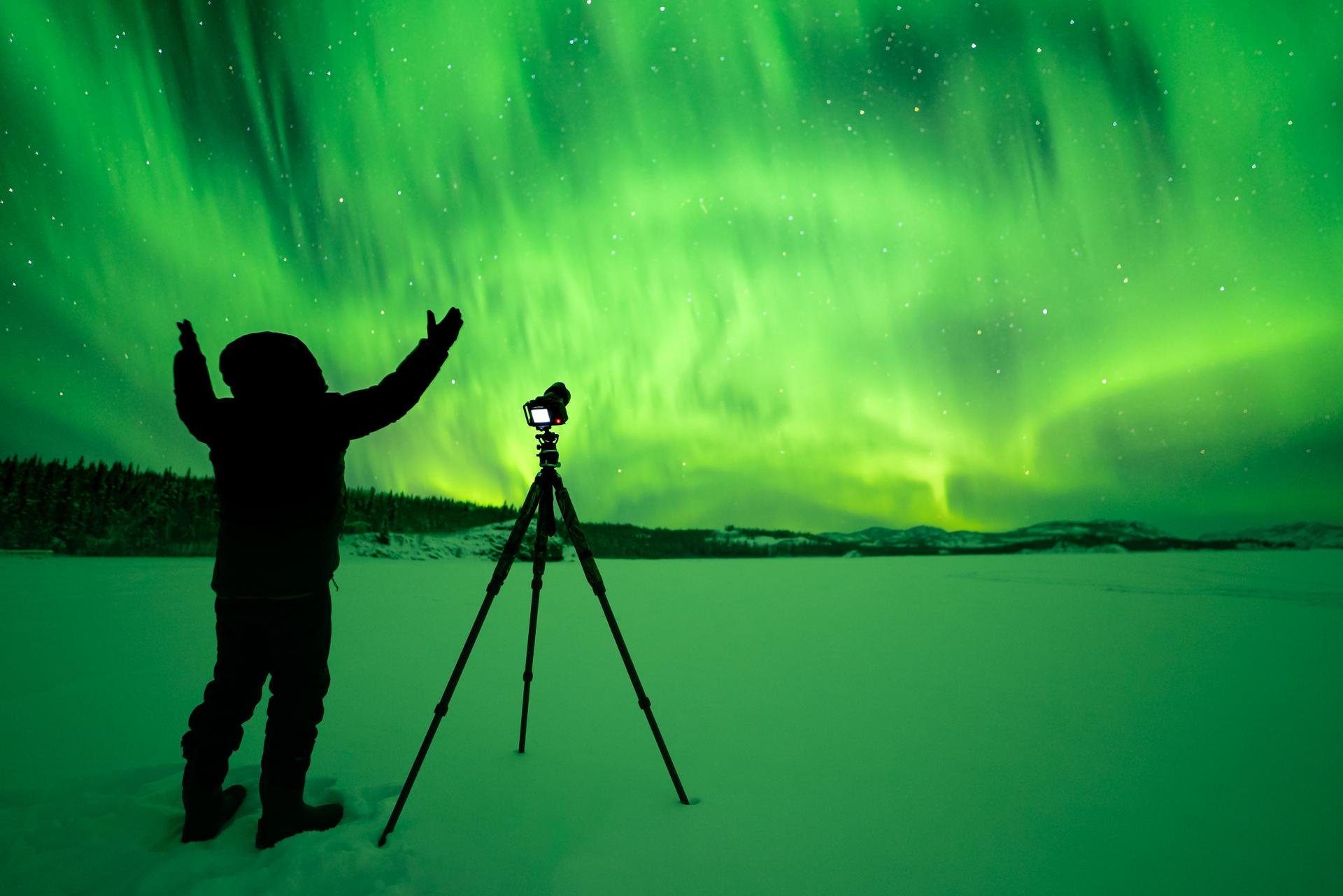 A person stands in the snow with arms raised, next to a camera on a tripod, capturing the vibrant green glow of the Northern Lights in the night sky.