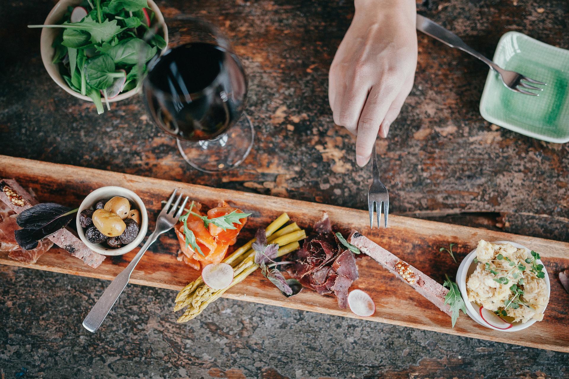 A person's hand reaching a fork towards a charcuterie board next to a small salad and a glass of red wine.