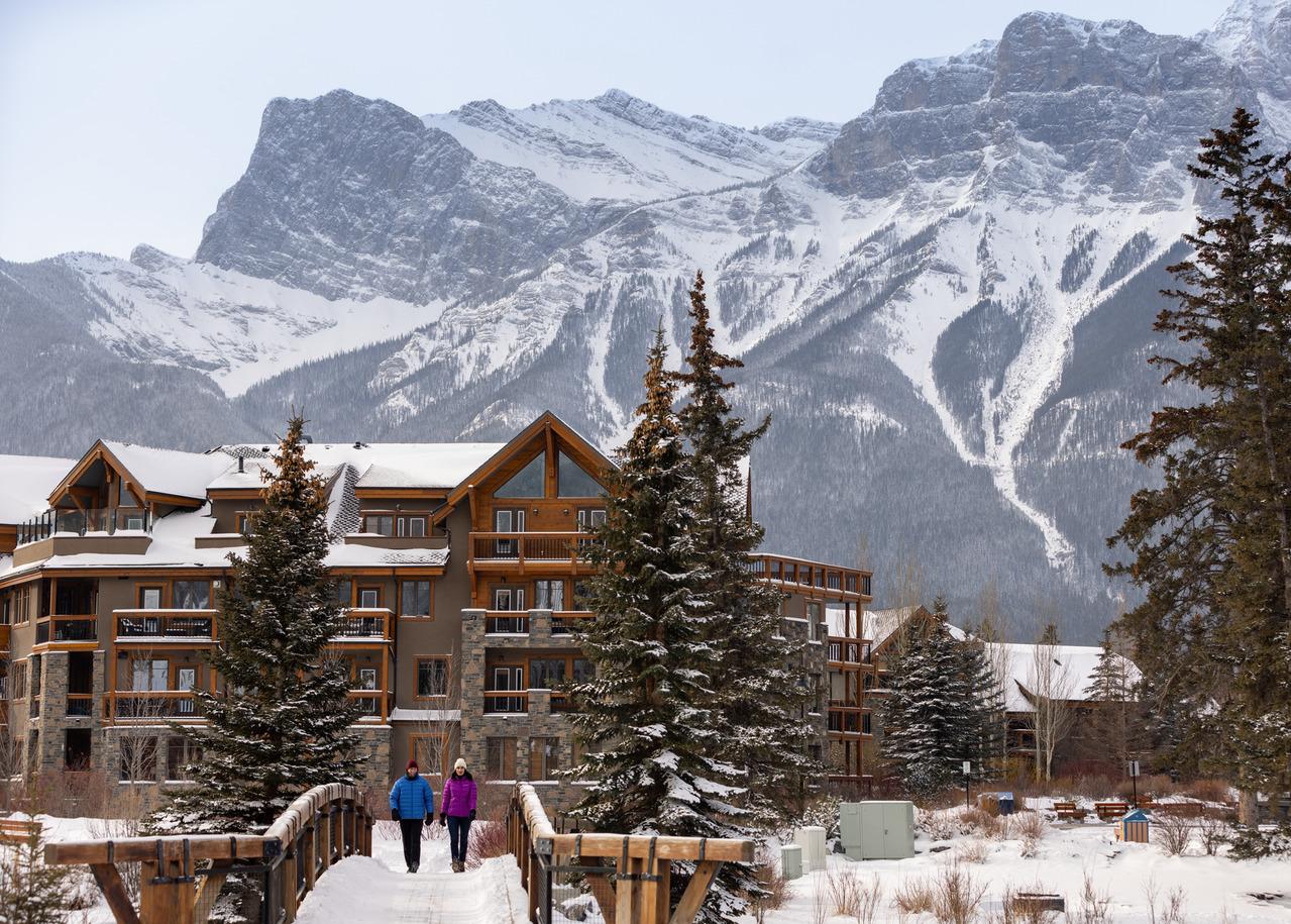 Two people walking on a snow-covered foot bridge toward the camera away from a hotel with the snowy Rocky Mountains in the background.