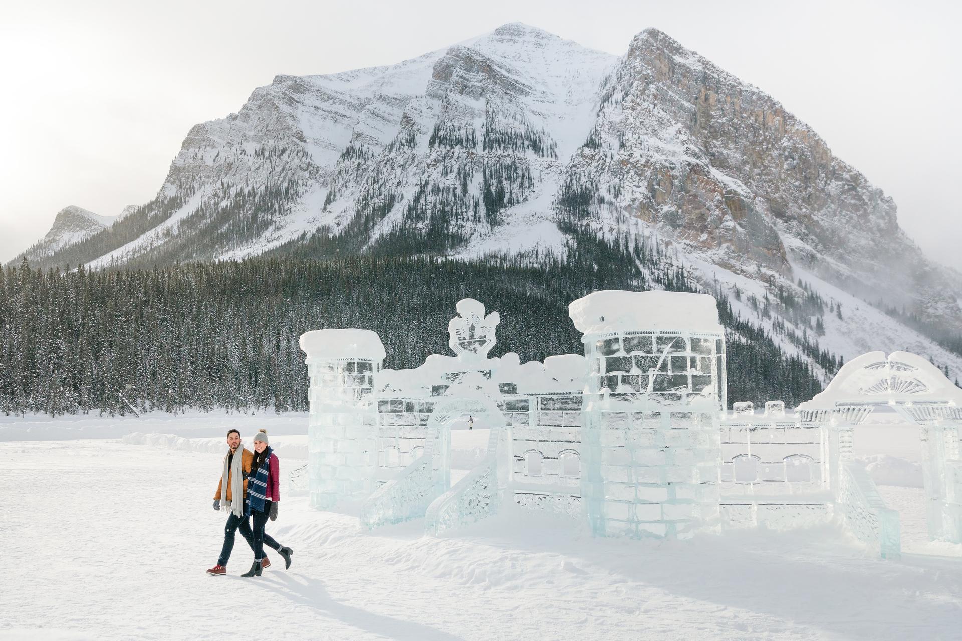 A man and a woman walking across the snow in front of an ice sculpture in the shape of a castle wall with a snowy mountain in the background.