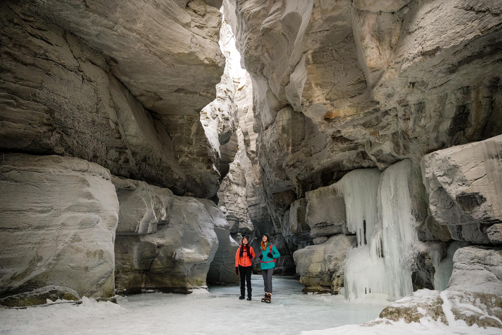 Two people standing on frozen ground inside a crevasse next to a frozen waterfall.