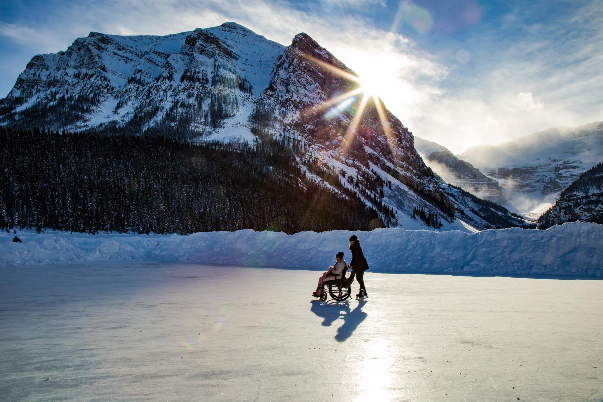 A person pushing another person in a wheelchair across a frozen lake in front of a snowy mountain.