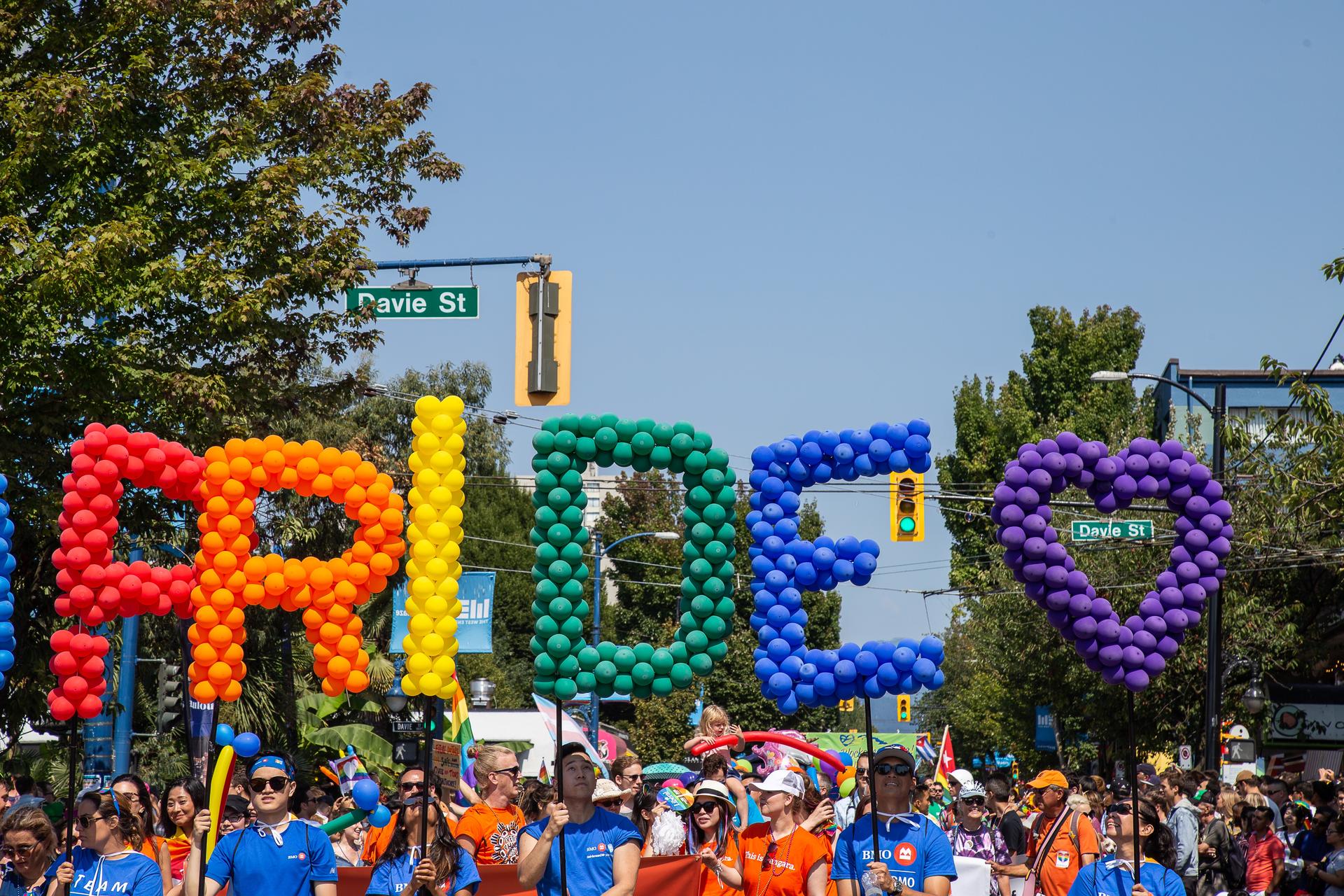 People marching down the street in Vancouver with balloons spelling Pride, Davie Street signs in the background.