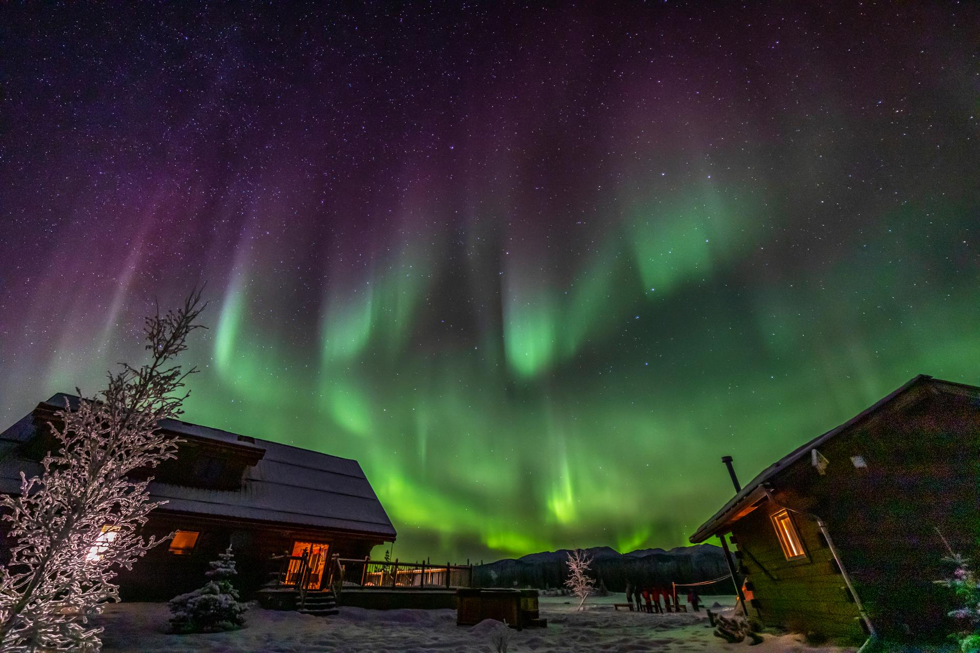 The northern lights above cabins in the snow.