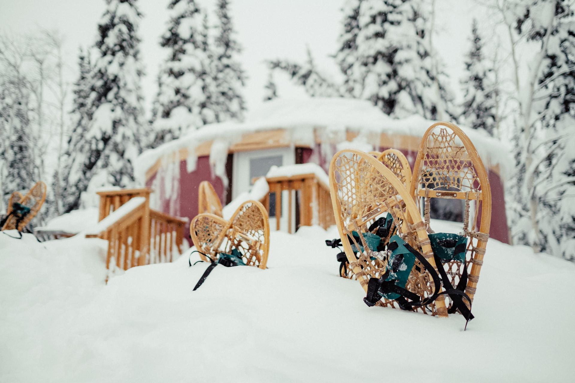 Pairs of snowshoes stuck upright in the snow in front of a snow-covered yurt in the forest.