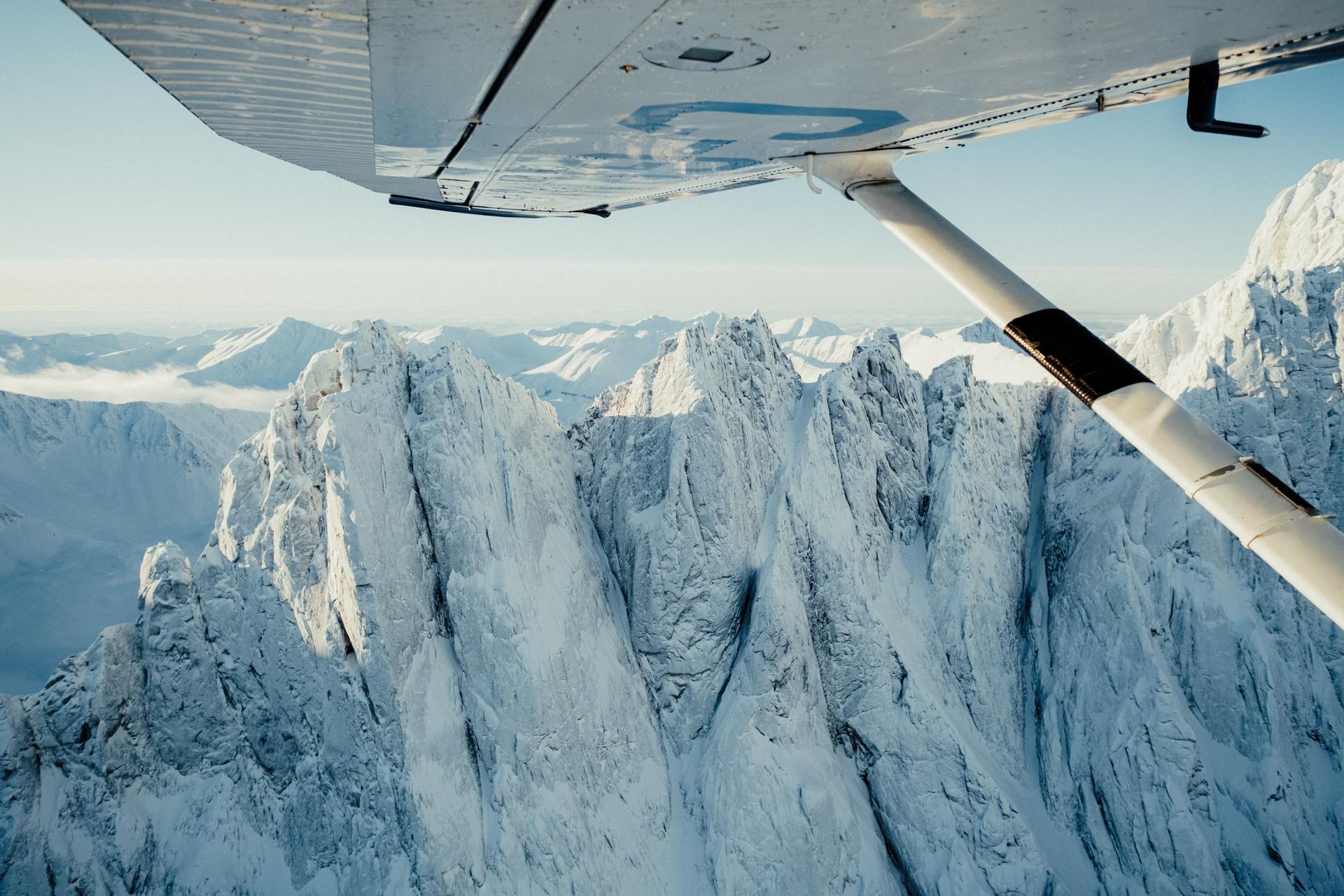 Craggy snow-capped peaks seen from beneath the wing of a prop plane.