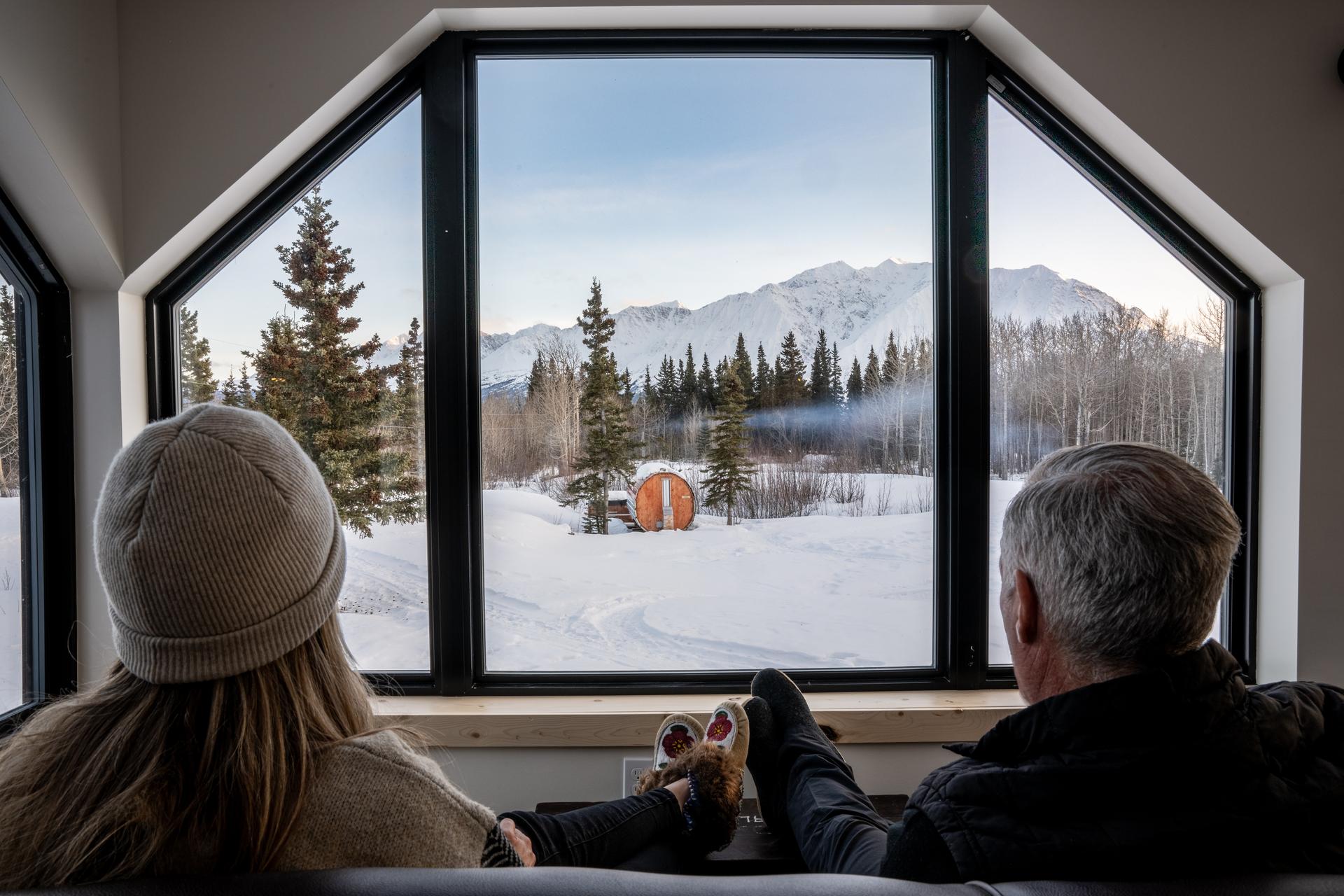 Two people relaxing on a sofa looking out a picture window at a snowy scene with forest and mountains in the background.