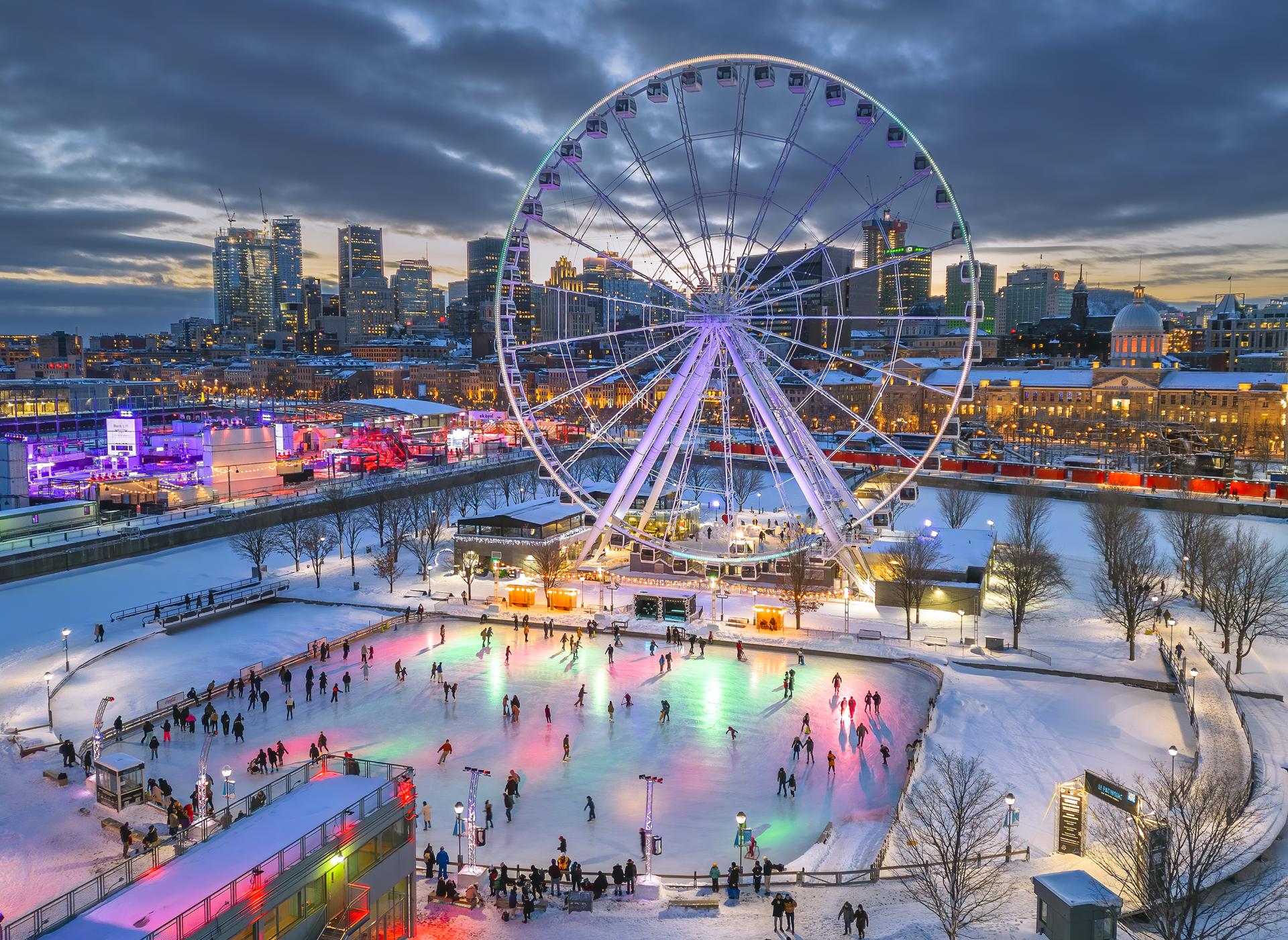 A large ferris wheel on the edge of an ice skating rink with downtown Montreal in the background at twilight.