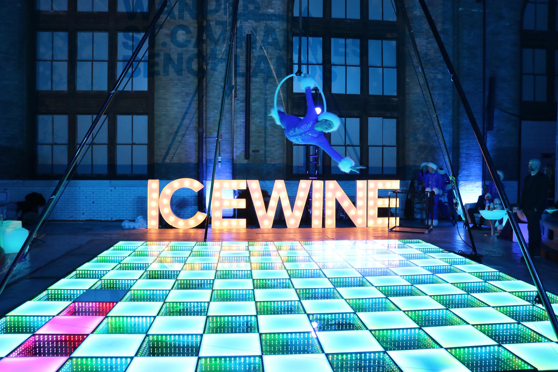 An acrobatic performer swinging on a hoop over a light-up checkerboard floor with the word ICEWINE spelled in lit up blocks at the edge.