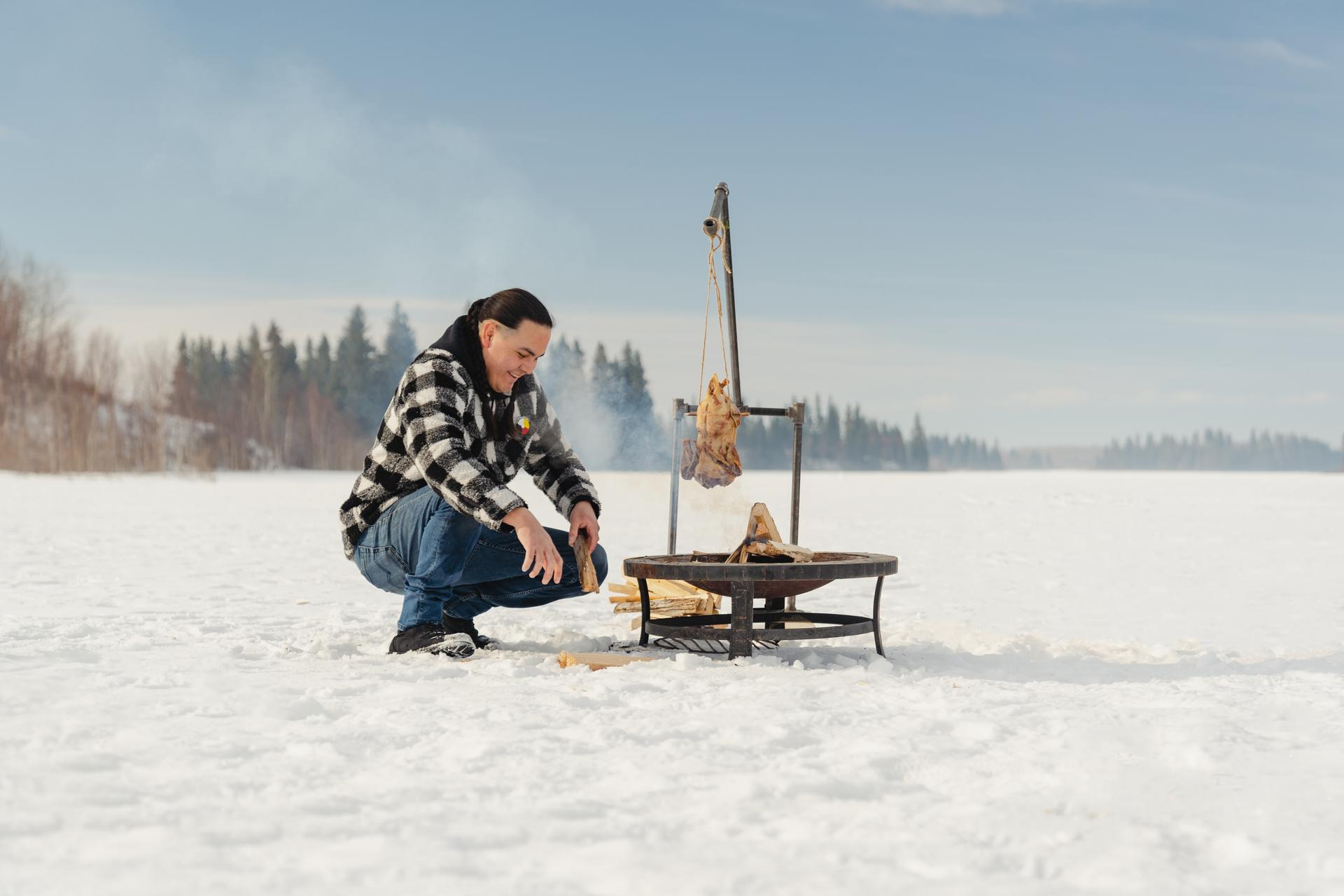 Person tending a fire in the snow with meat hanging to cook