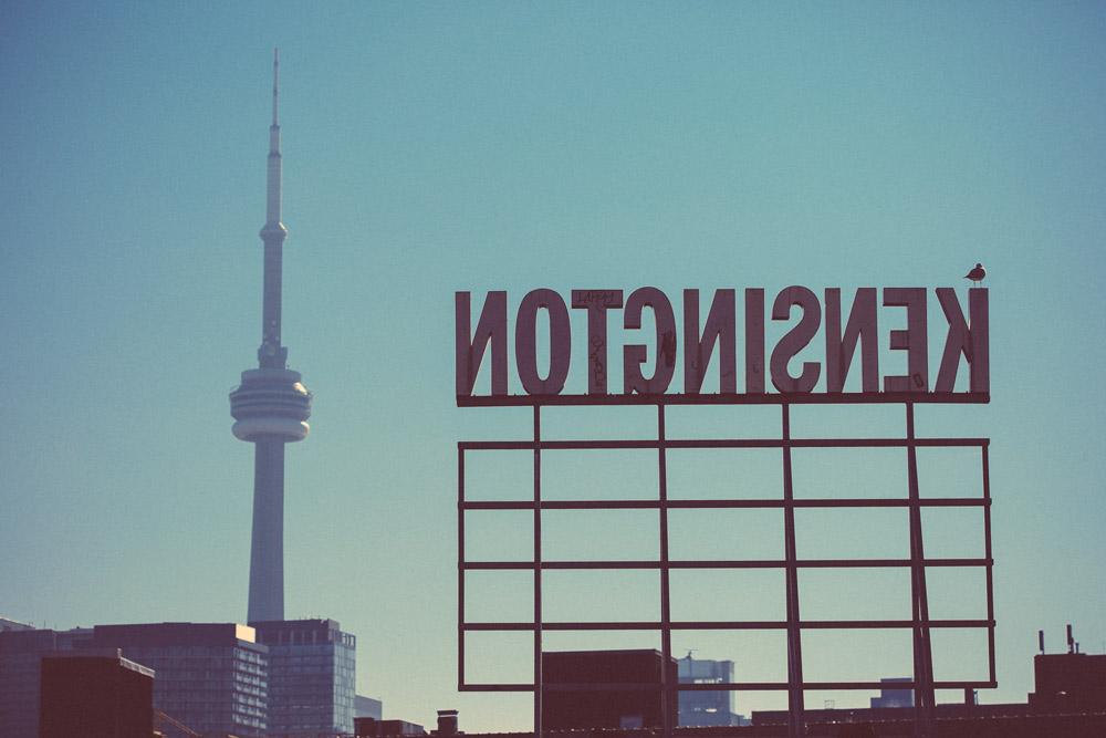 The Kensington Market sign in Toronto seen from behind with the CN Tower in the background.