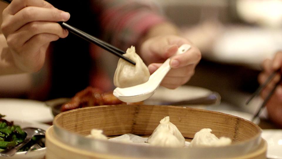 A person lifting a soup dumpling with chopsticks over a steaming bamboo basket