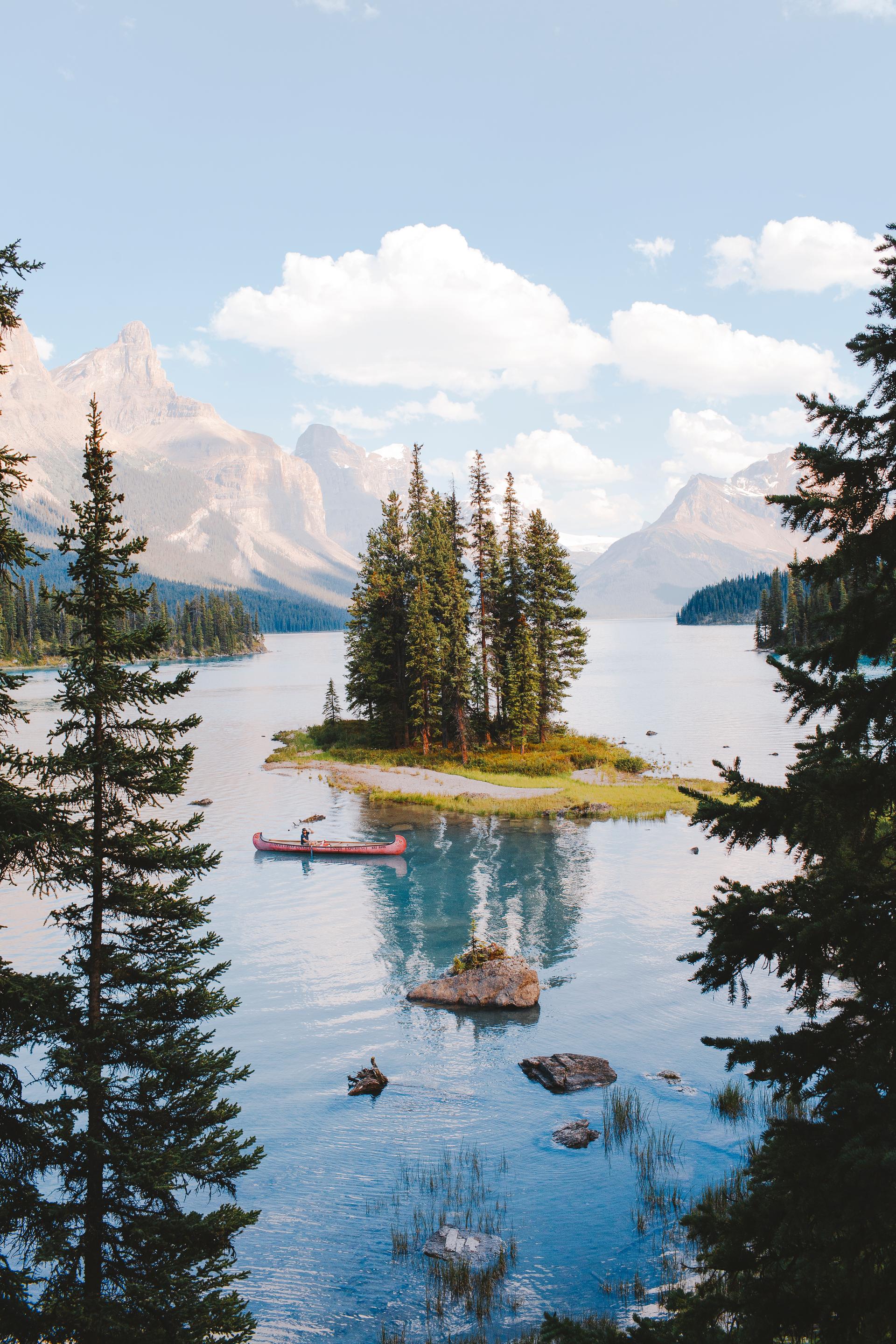 Person paddles a red canoe near a small forested island on a calm mountain lake
