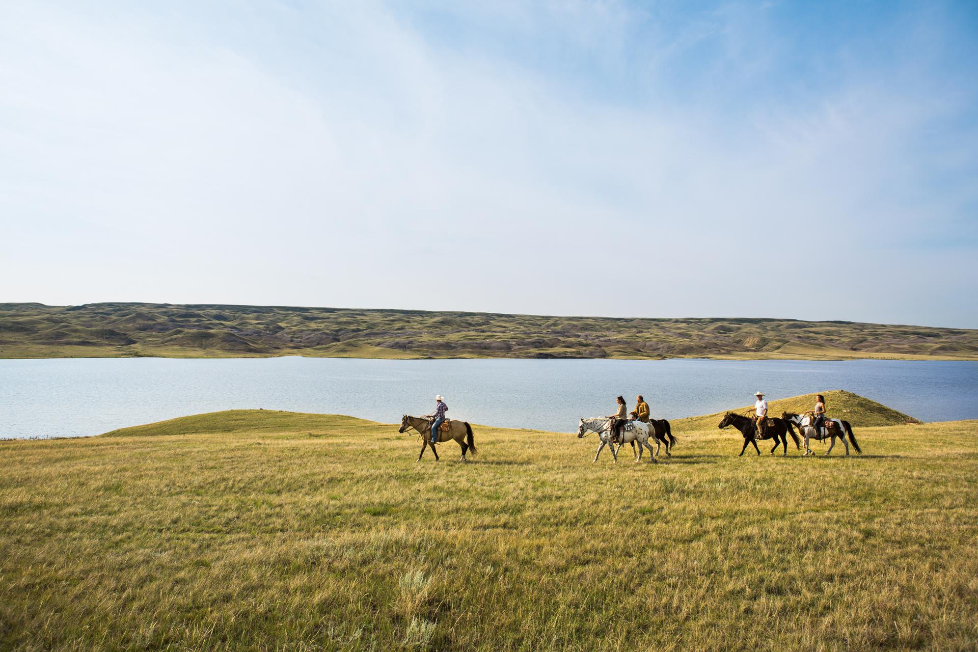 Group of people ride horses across grassy plains beside a lake under a clear blue sky