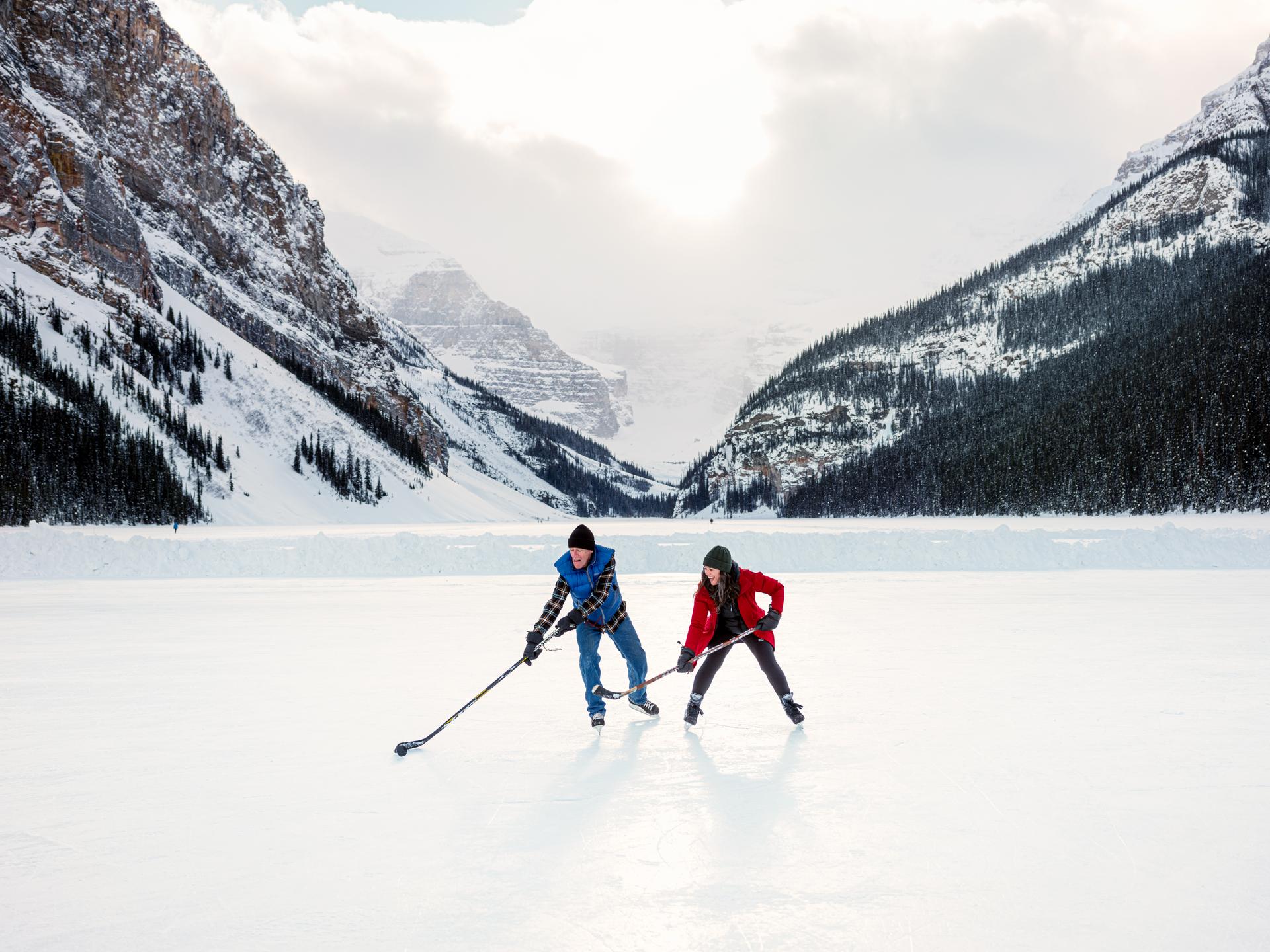 Two people playing ice hockey on a frozen lake surrounded by snow-covered mountains