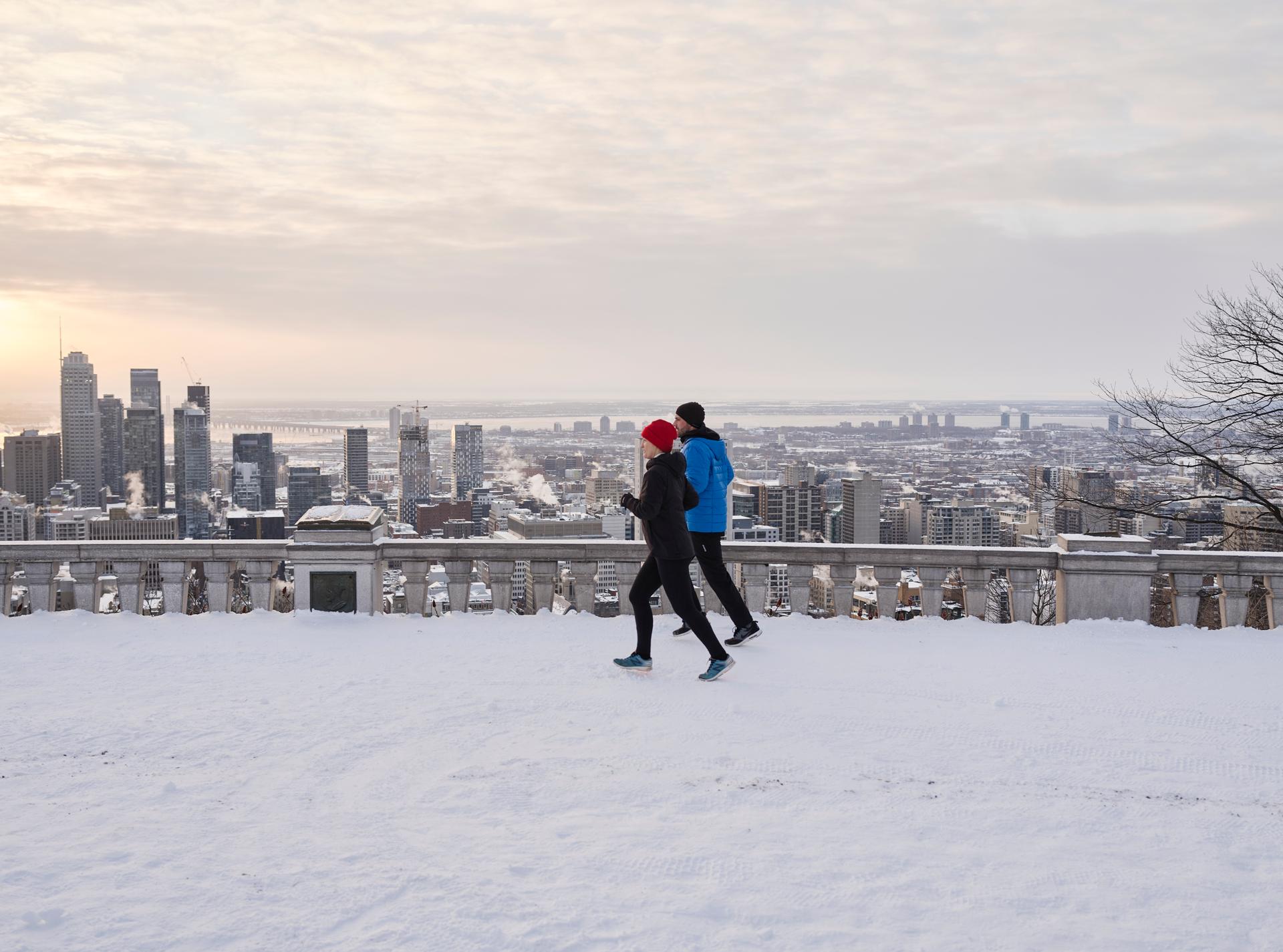 Two people jogging in winter on a snowy lookout with a view of downtown Montreal’s skyline