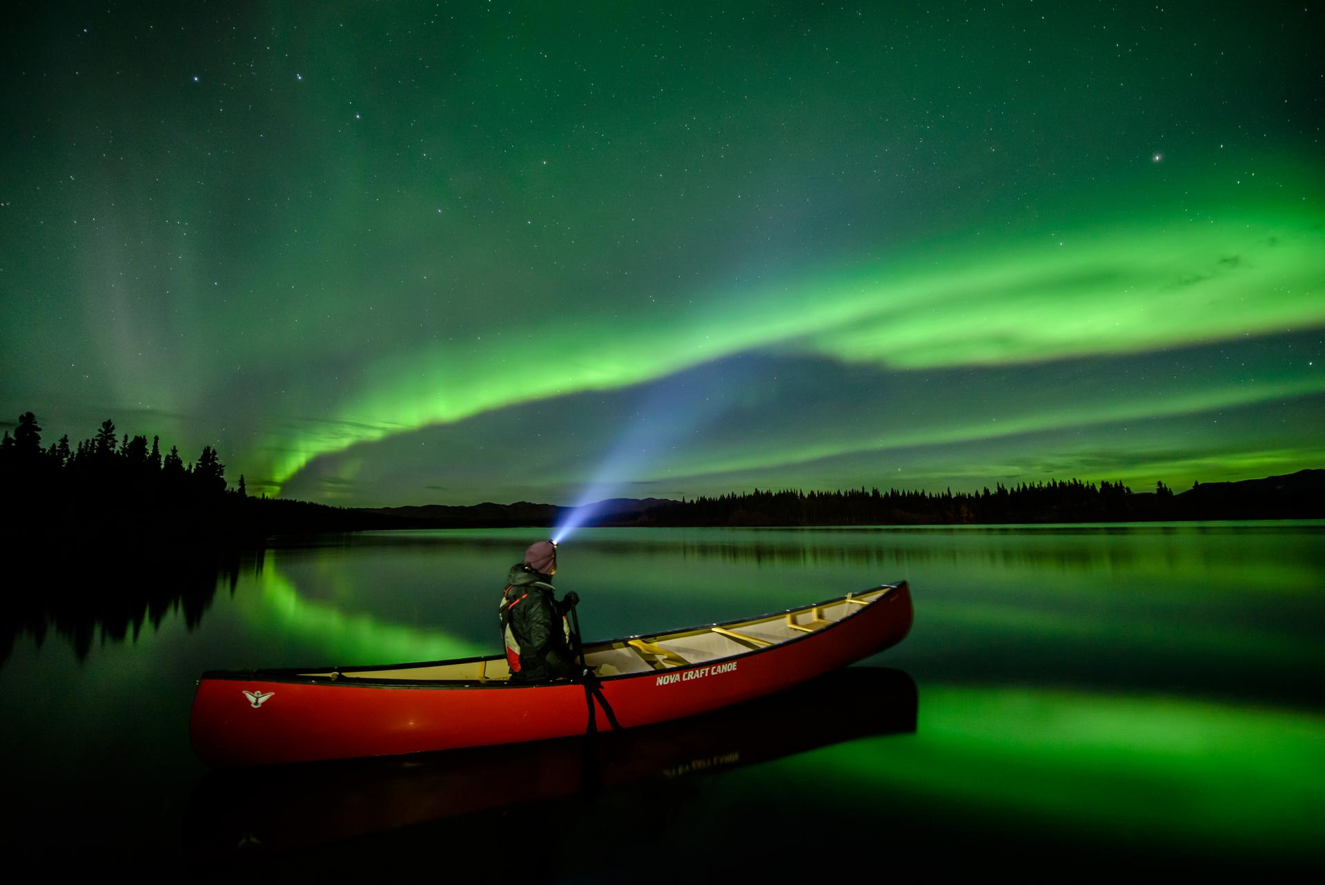 A person in a red canoe watches green northern lights reflected on a calm lake at night