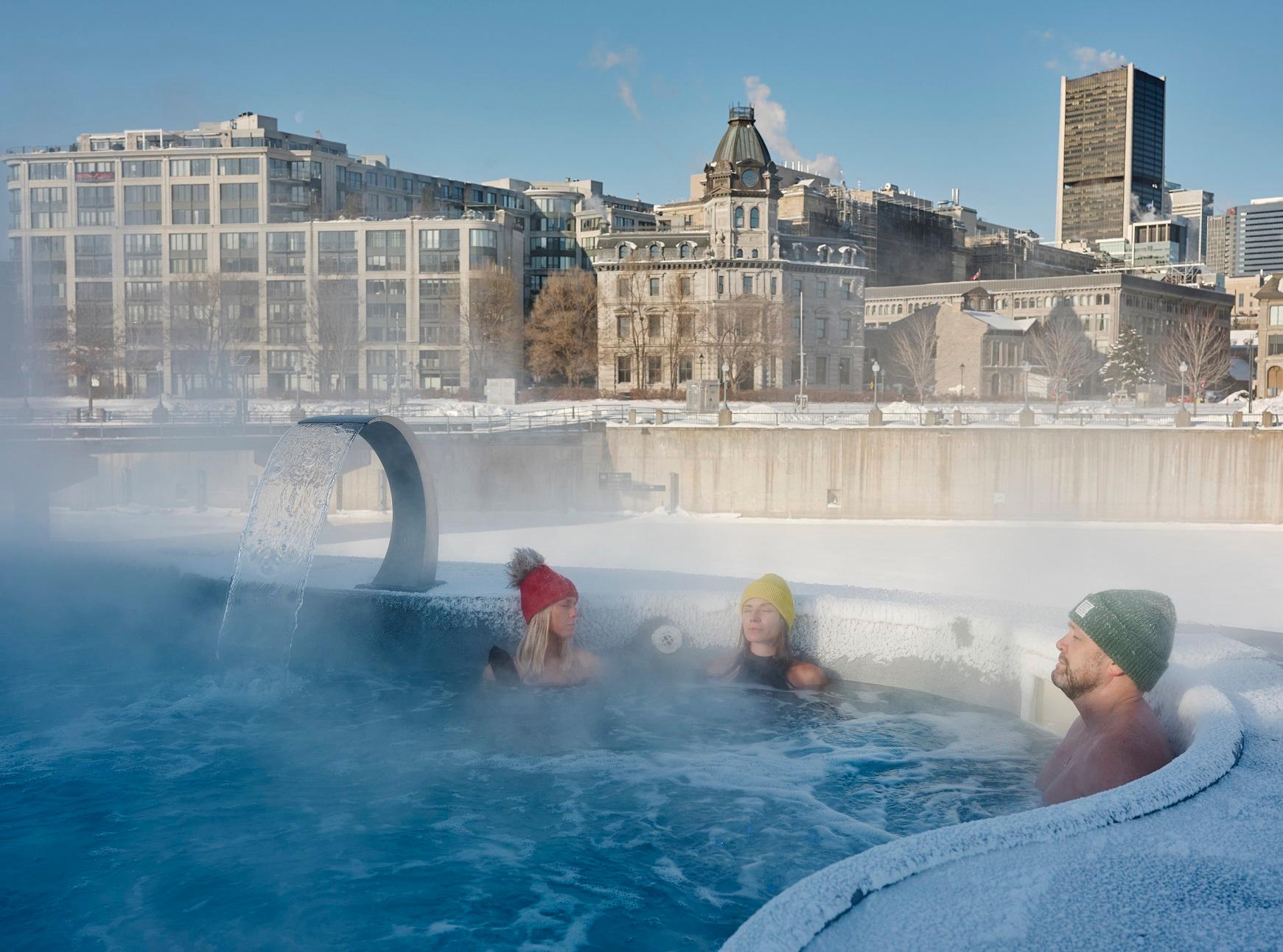 Three people relaxing in a steaming outdoor spa overlooking a winter cityscape