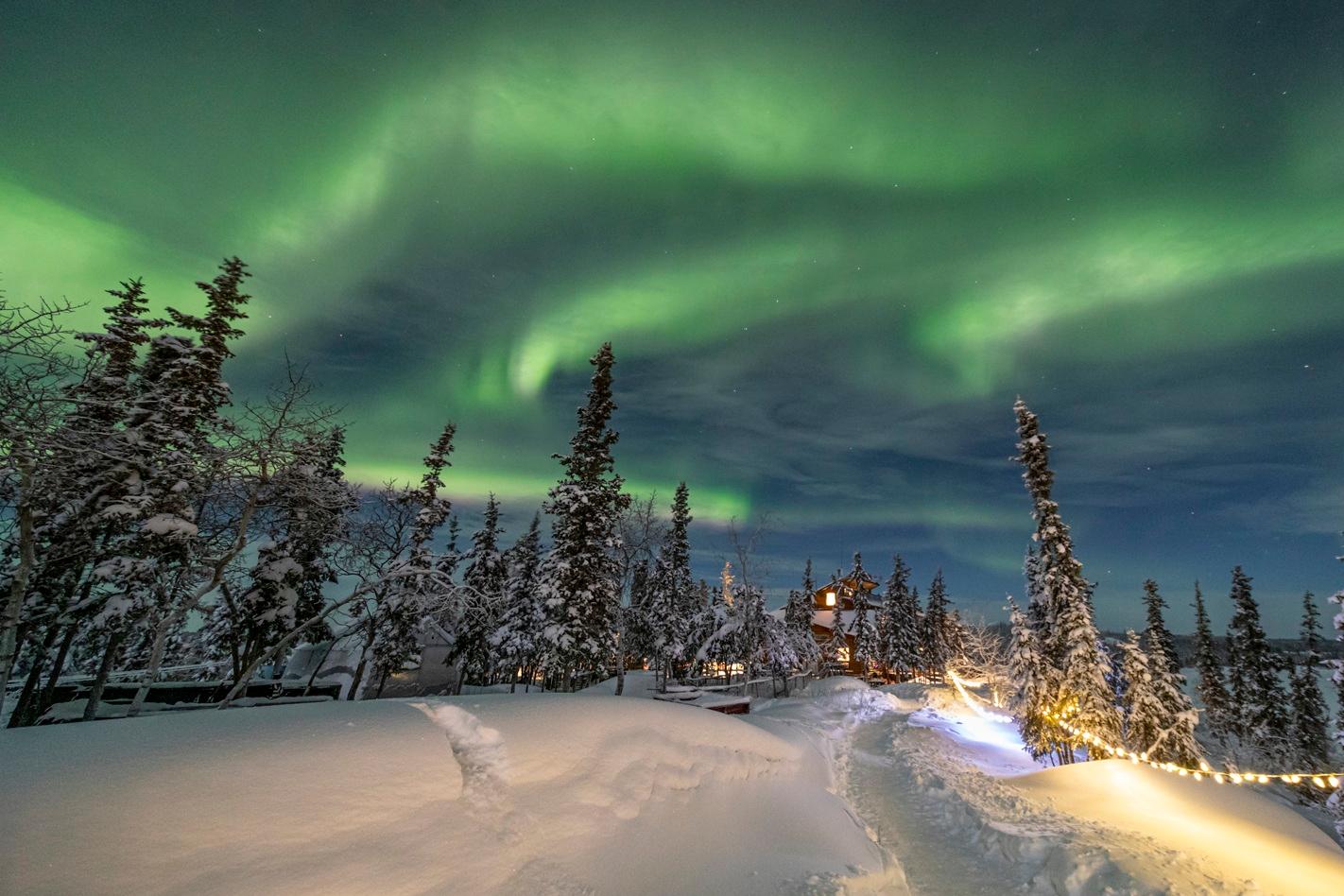Green northern lights above a snow-covered forest and illuminated lodge