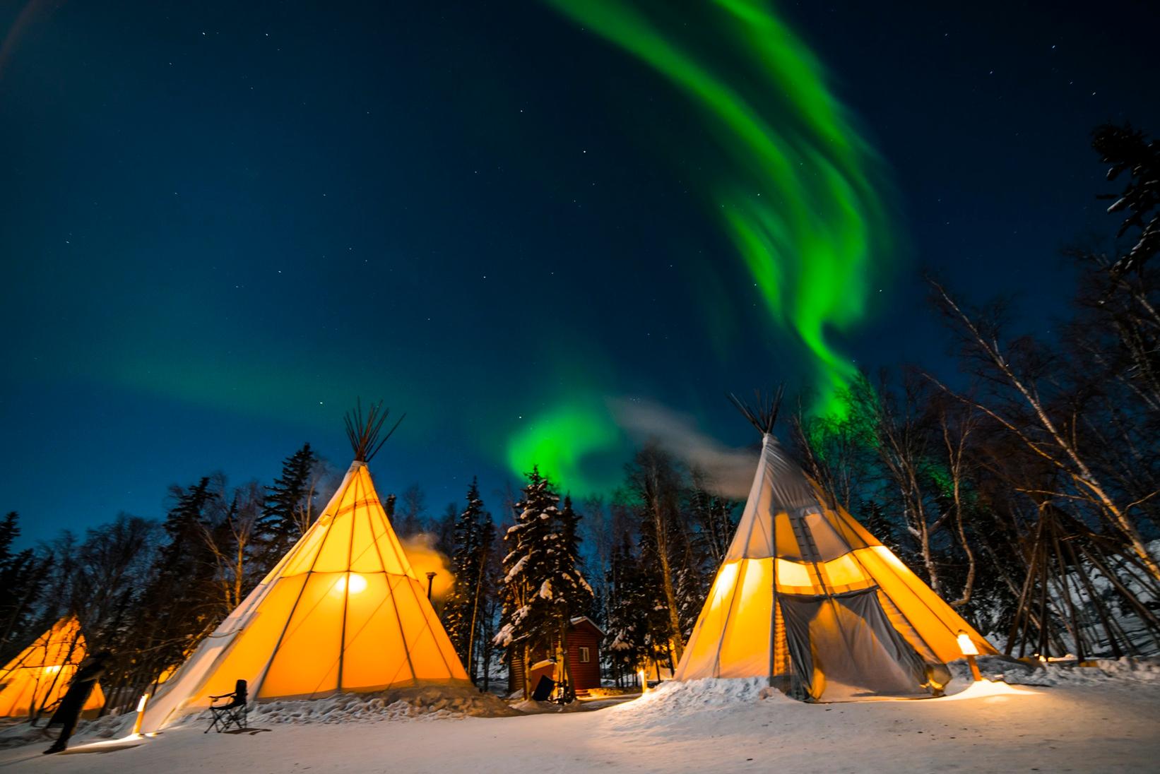 Illuminated teepees beneath green northern lights in a snowy forest