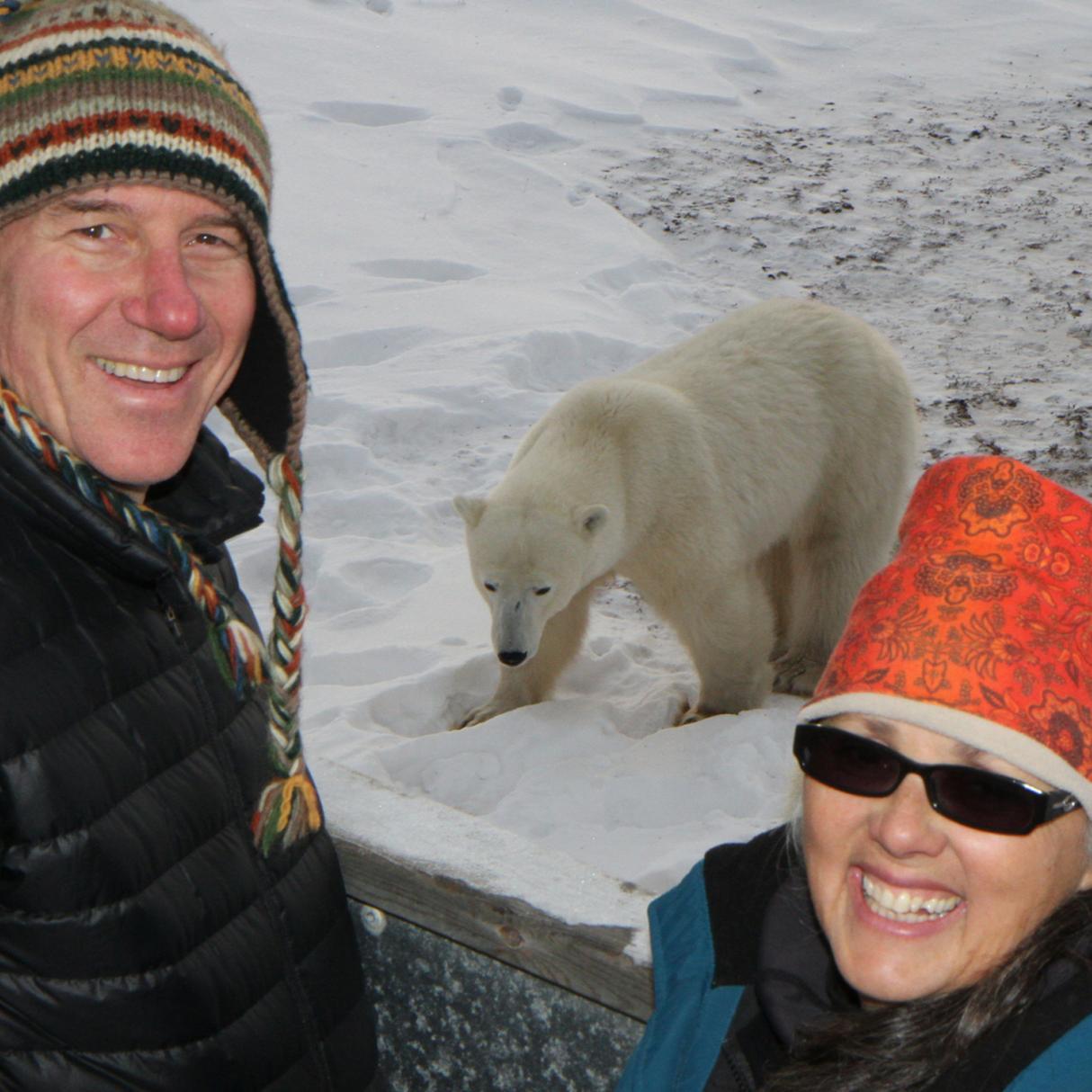 Two people who wear an orange and a striped hat stand smiling in front of a nearby polar bear walking through the snow. 
