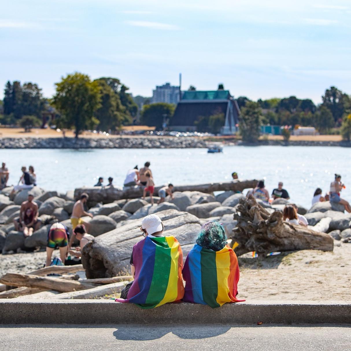 Two beachgoers wrapped in pride flags sit on the shore together