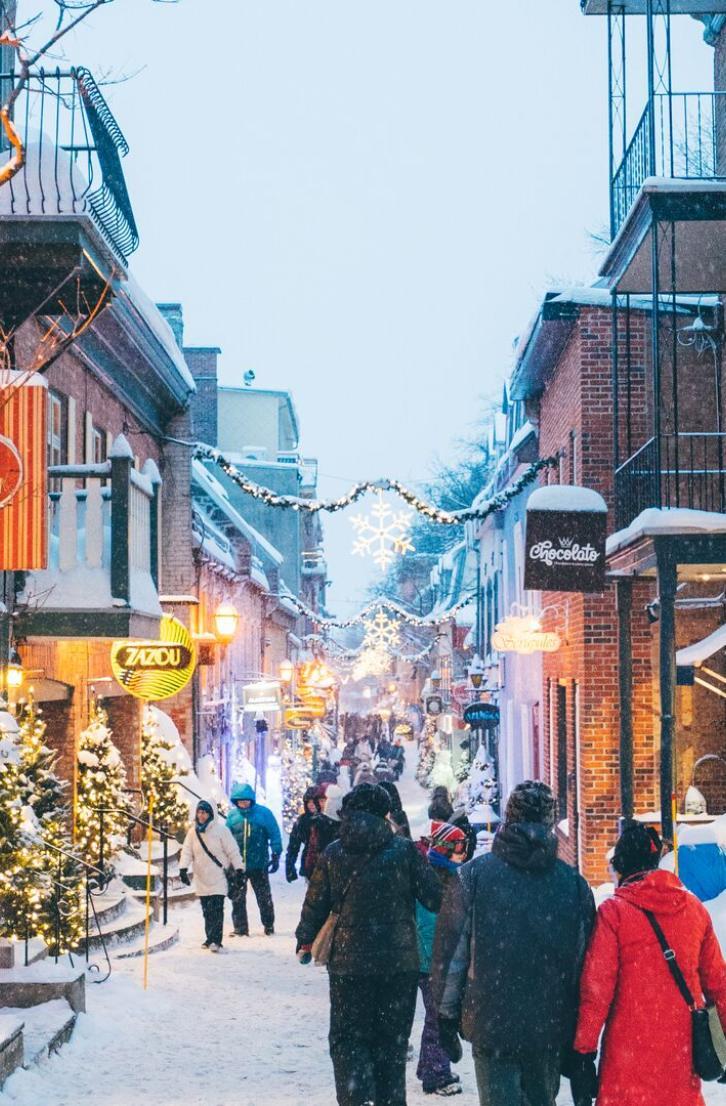 People stroll through a charming snow-covered street lined with illuminated shops and decorated with festive lights.