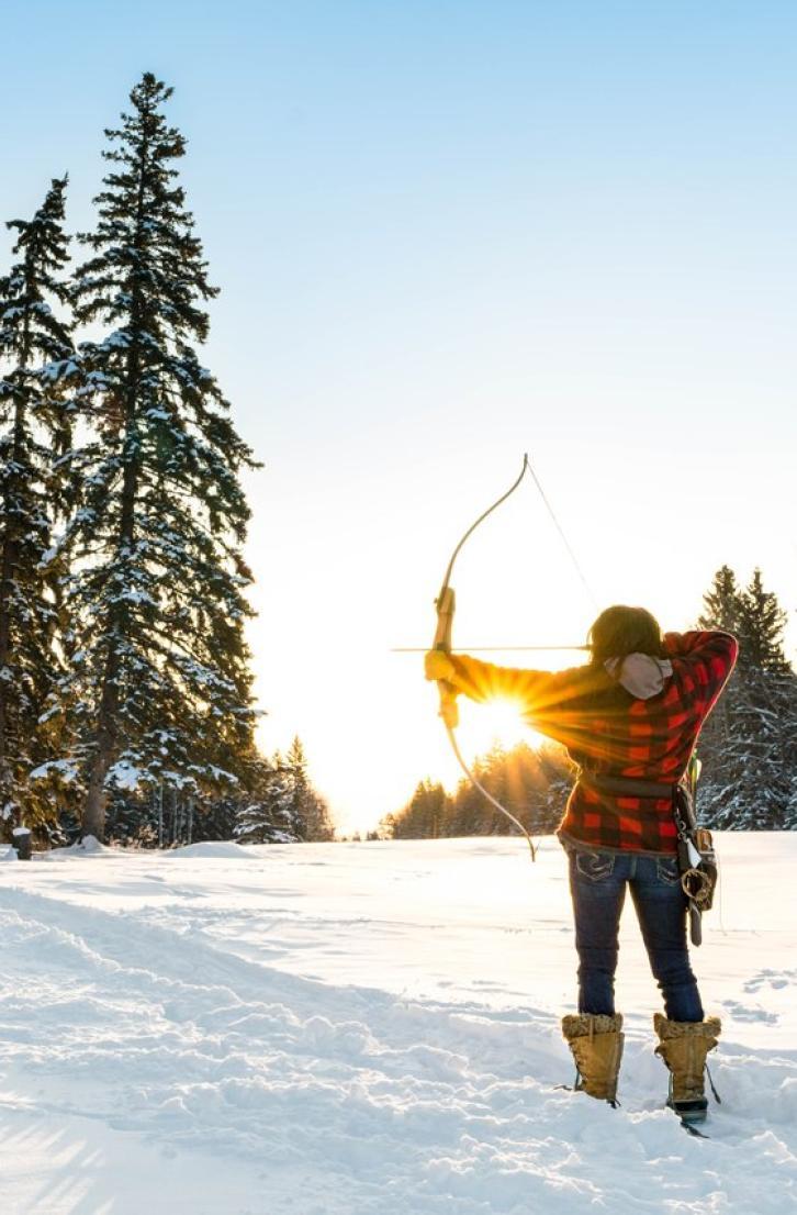 A person practices archery in a snowy forest clearing at sunset.