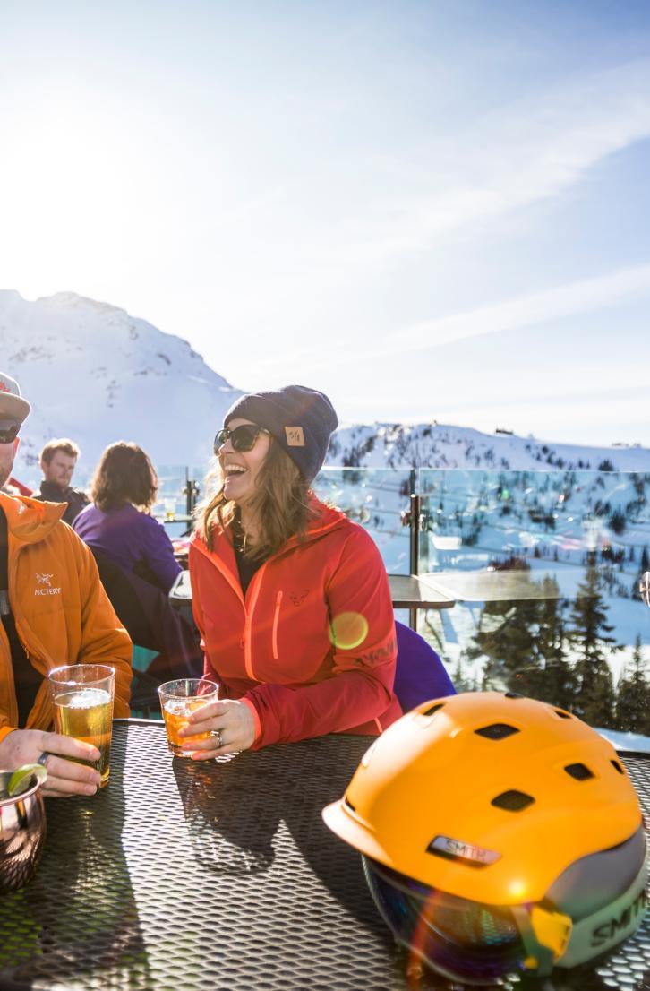 A group of friends enjoys drinks at a sunny mountain terrace with snow-covered slopes and ski lifts in the background.