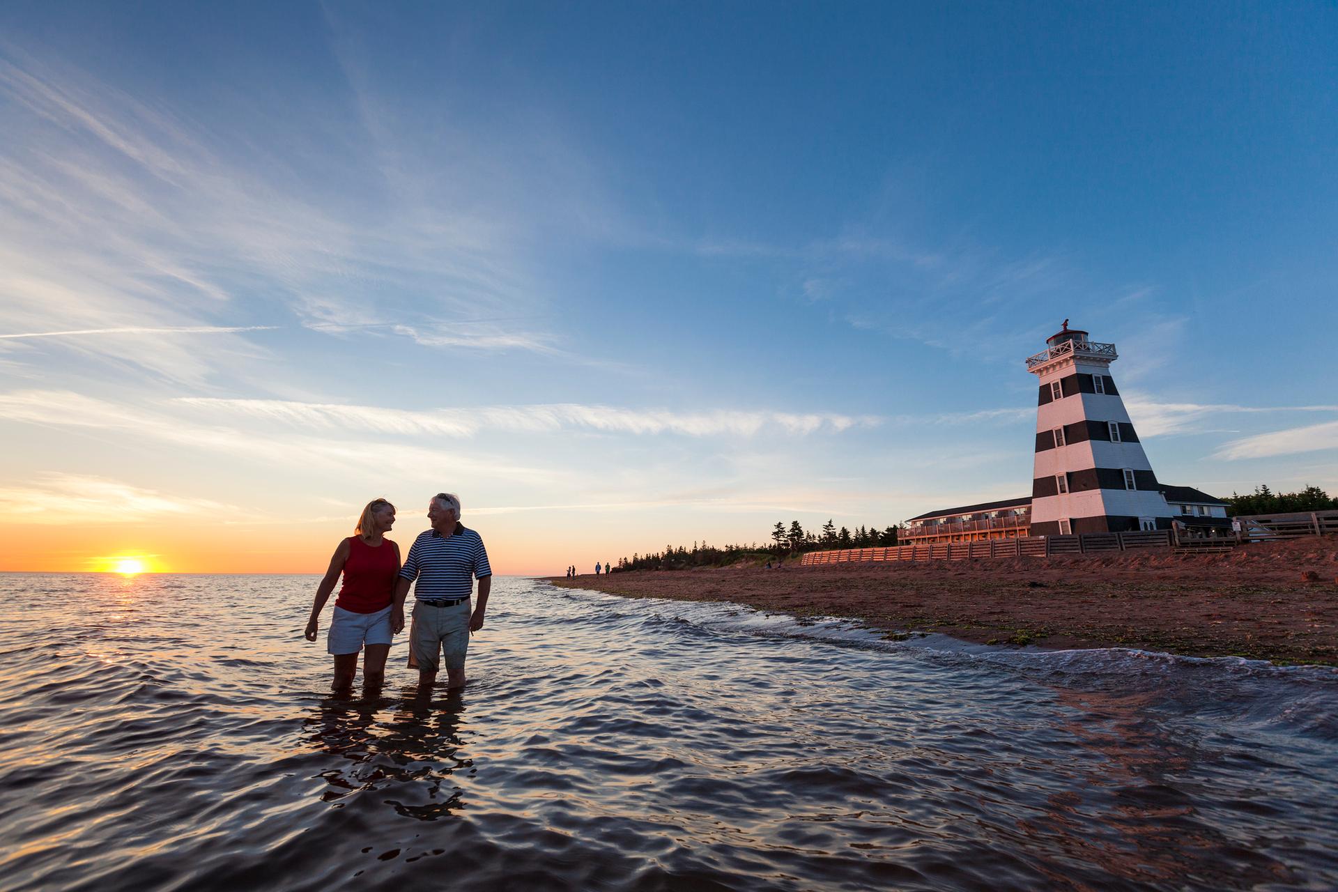 Two people walk in the waters at a Prince Edward Island beach, in view of a lighthouse