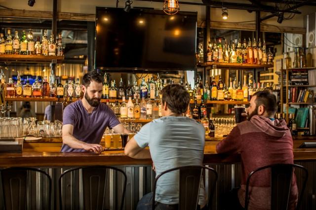 Two patrons enjoy a beer at a bar, alongside a bartender