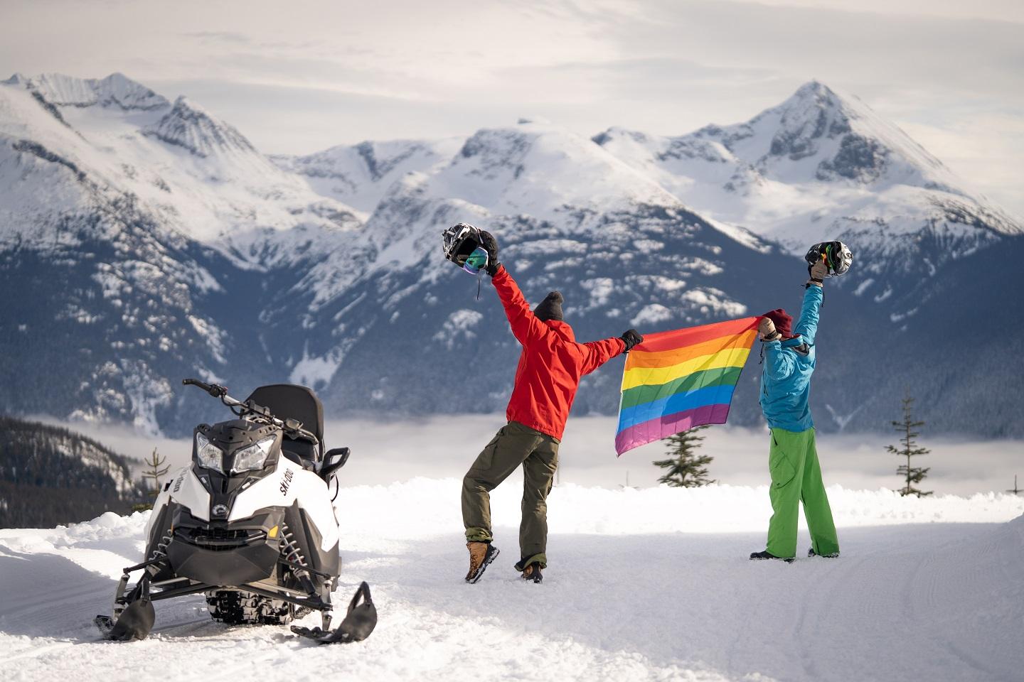 Two skiers hold up a pride flag next to a snowmobile in the mountains of Whistler