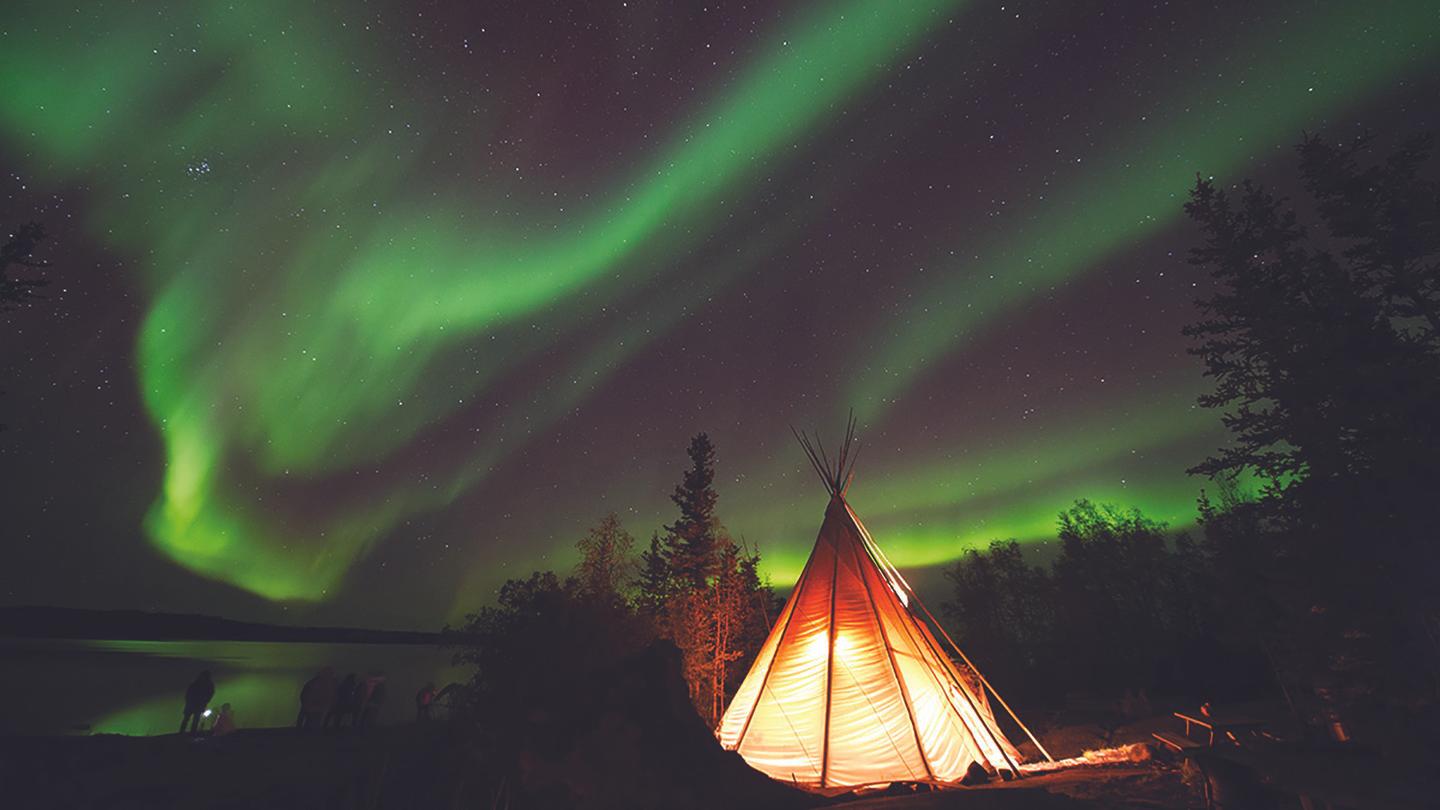 Teepees along the river under vibrant green northern lights