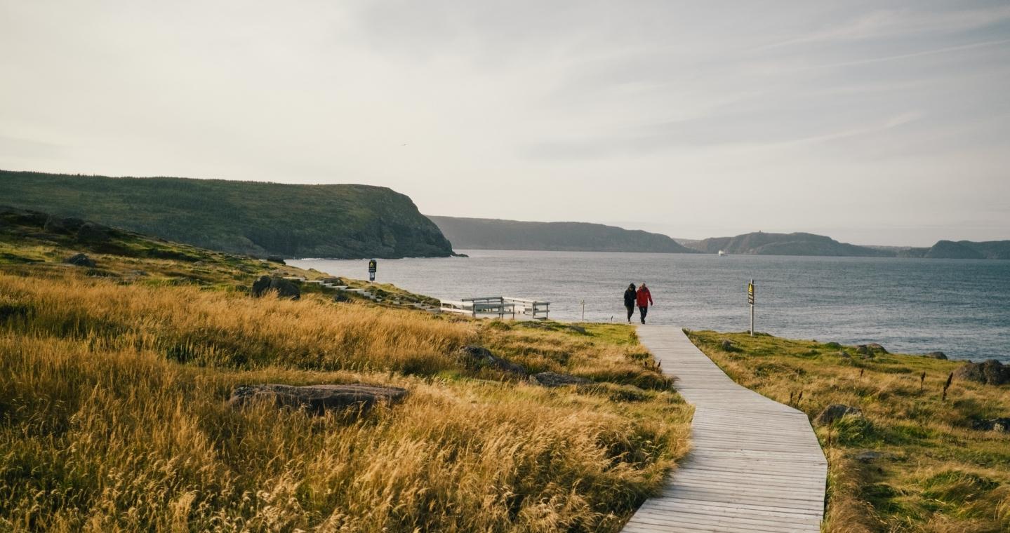Two hikers walk a boardwalk adjacent to a grassy seaside cliff