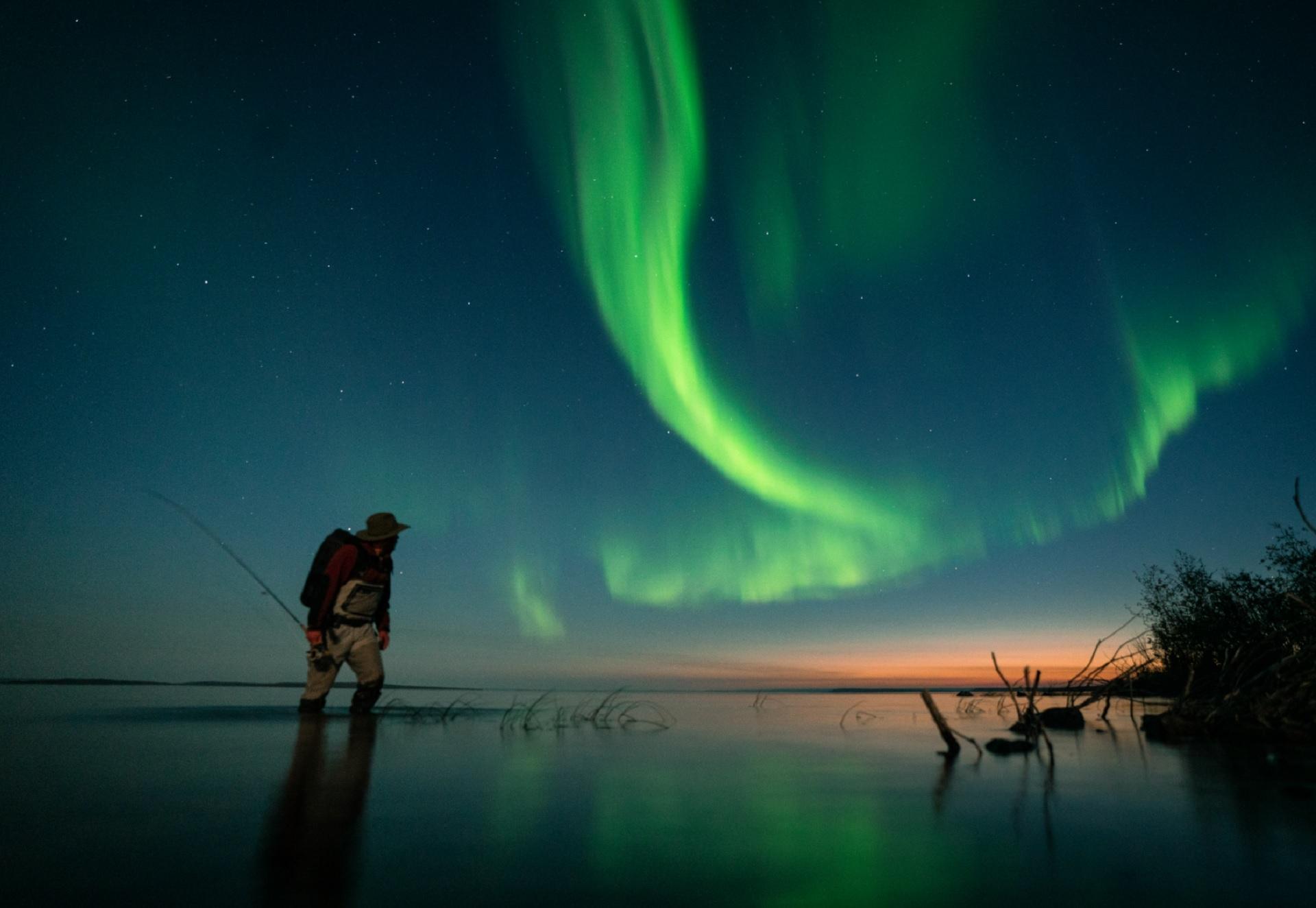 Person standing with fishing rod in the lake looking at the northern lights