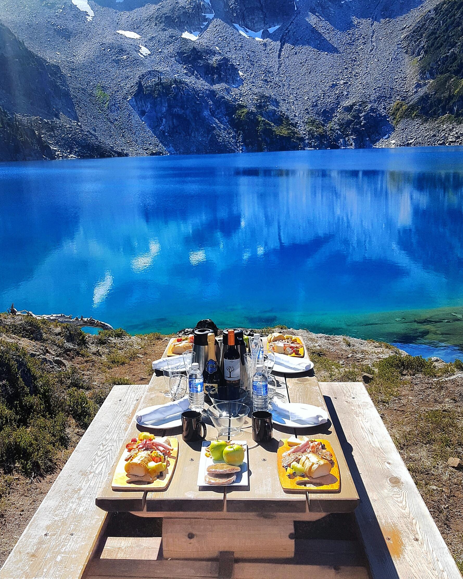 An elegant meal laid out on a picnic table, next to a blue lake at the bottom of a mountain