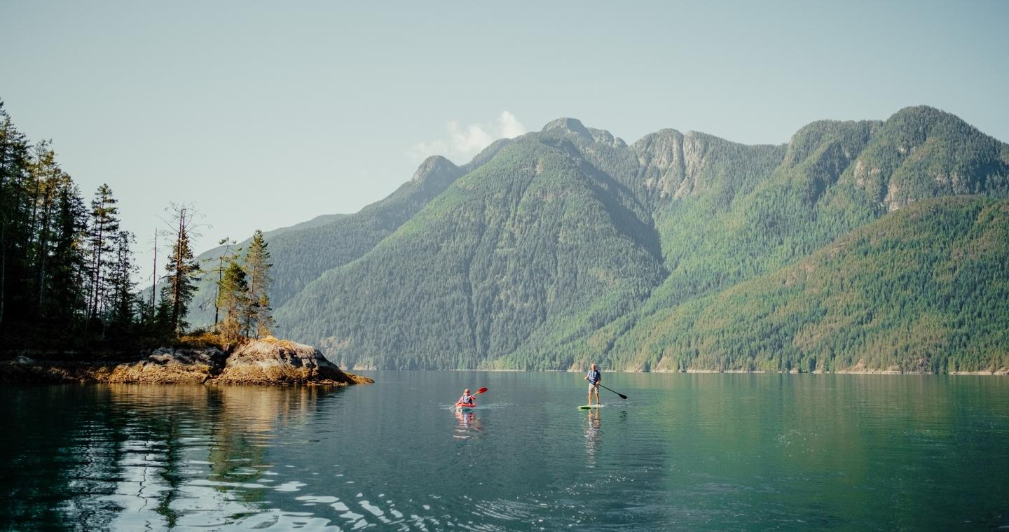 Two people paddle a canoe and a paddleboard on a lake at the foot of a mountain